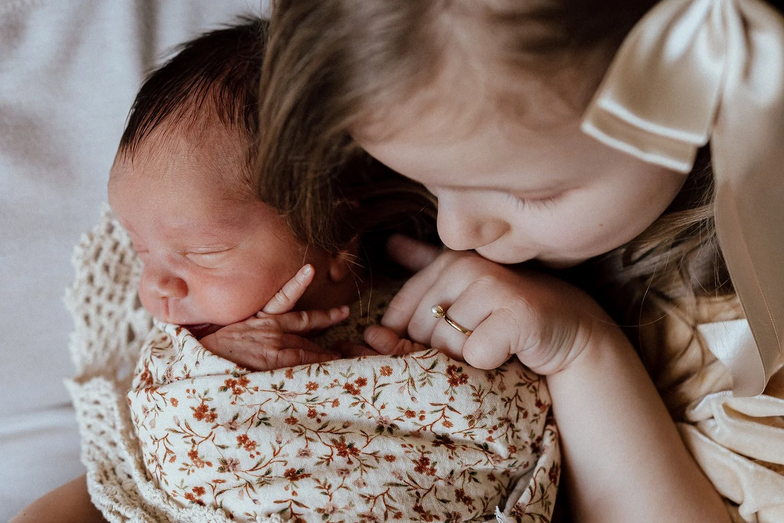 A young girl with blonde hair gently kisses a newborn baby resting on a floral blanket. The baby is wrapped in a crochet blanket and has a peaceful expression.