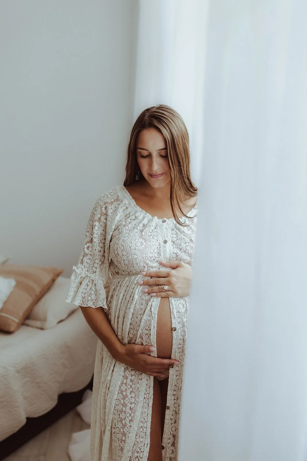 A pregnant woman in a lace dress gently touching her belly with her eyes closed, standing near a sheer curtain in a softly lit room.
