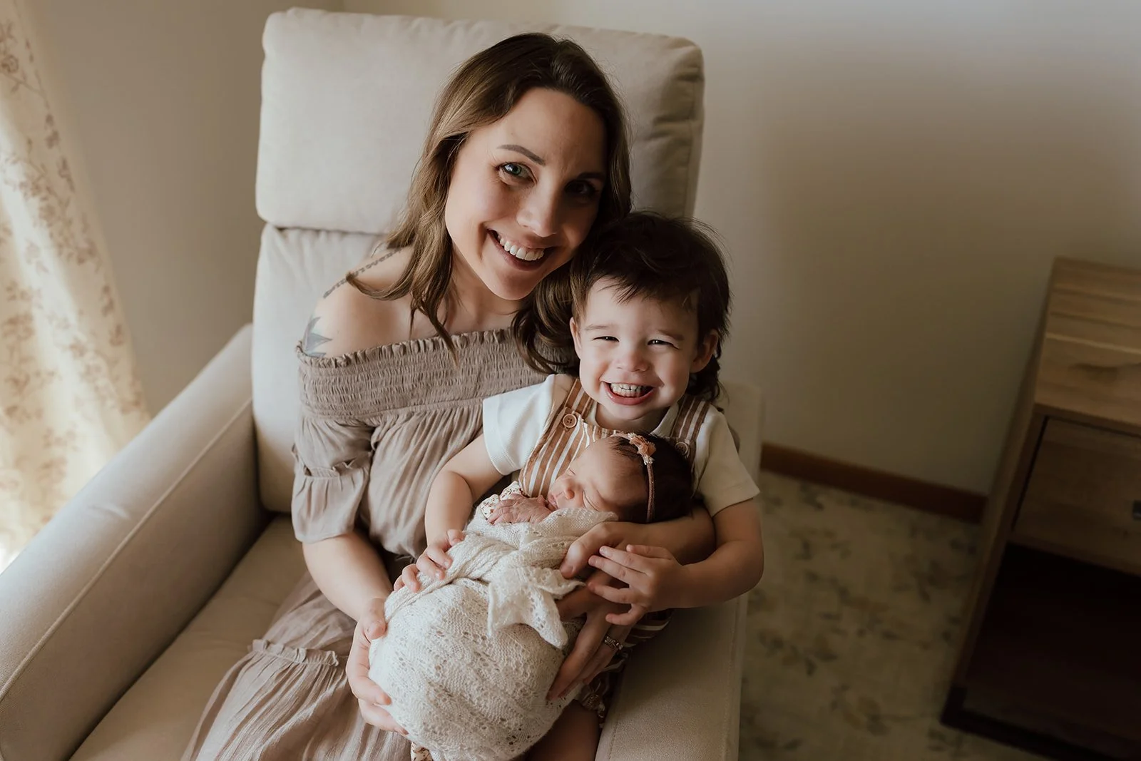 A woman sitting on a beige couch holding a newborn baby wrapped in a cream blanket, with a young girl smiling and holding the baby.
