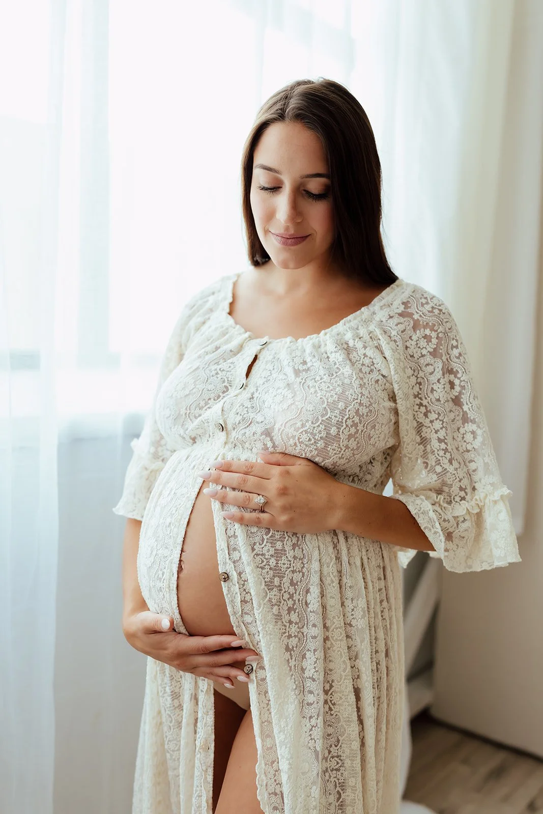 Pregnant woman in a cream lace dress standing indoors, gently holding her baby bump with her eyes closed.