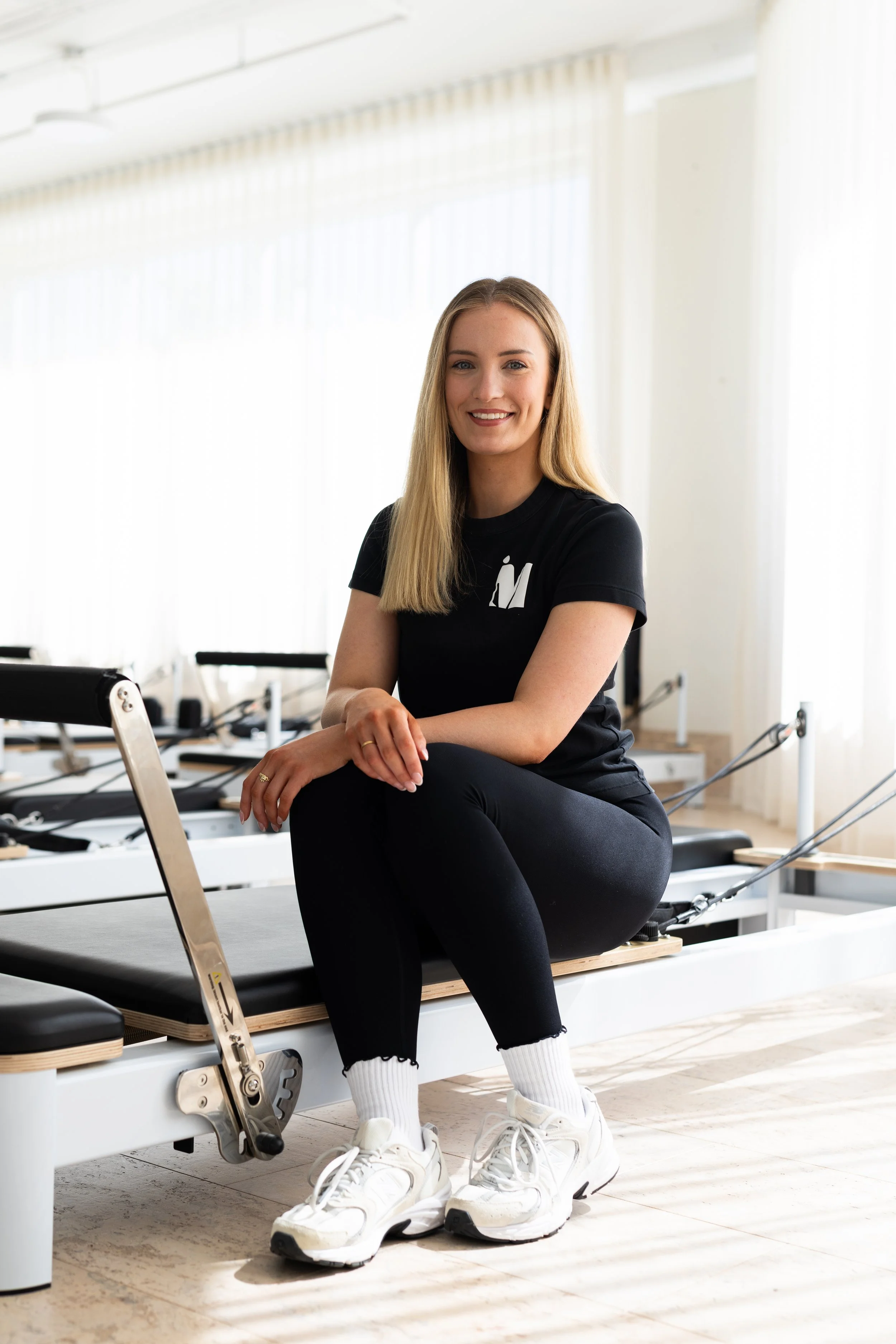 A woman standing in a Pilates studio, smiling, holding an exercise ball, surrounded by Pilates equipment such as reformers and mirrors.