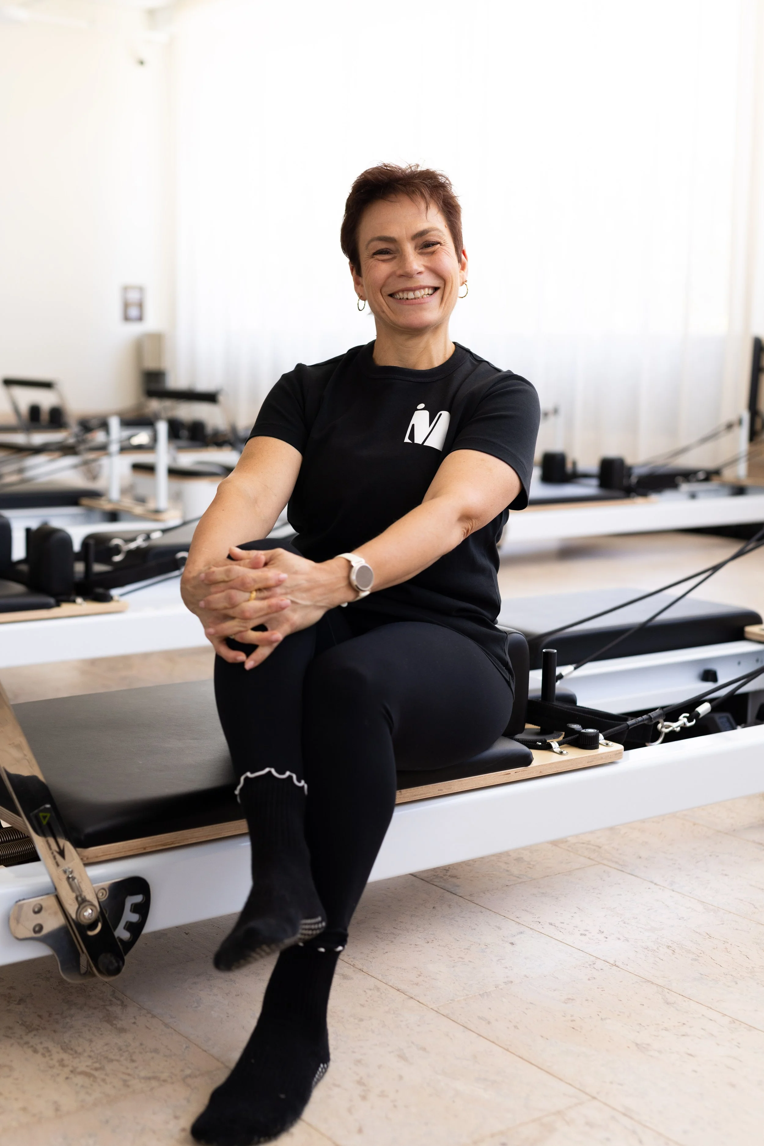 A woman doing Pilates on a reformer machine in a fitness studio with mirrors and plants.