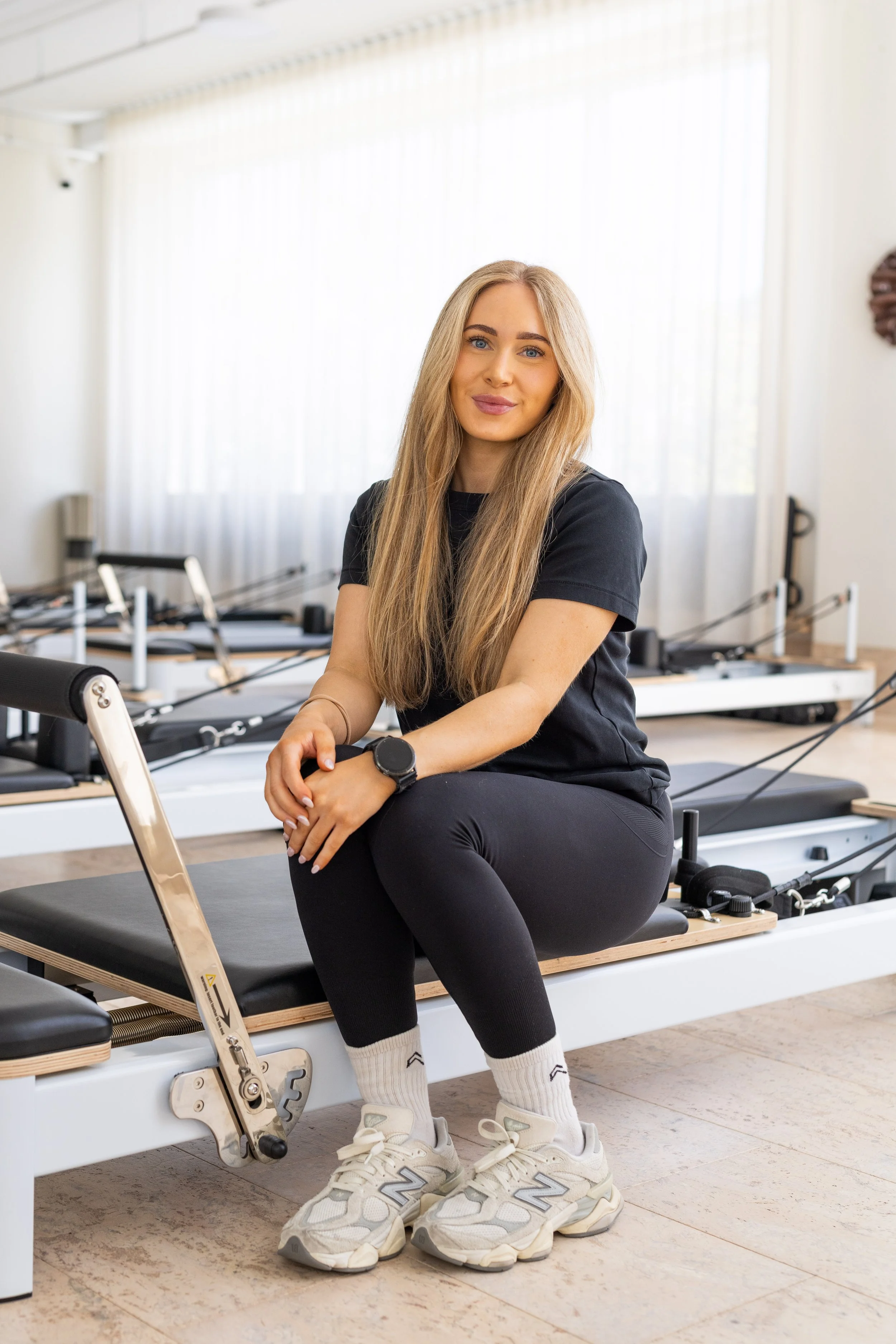 A woman with brown hair sitting on a Pilates reformer machine in a fitness studio, smiling at the camera. She is wearing a black t-shirt with a white logo and black leggings, with one foot in a white sock.