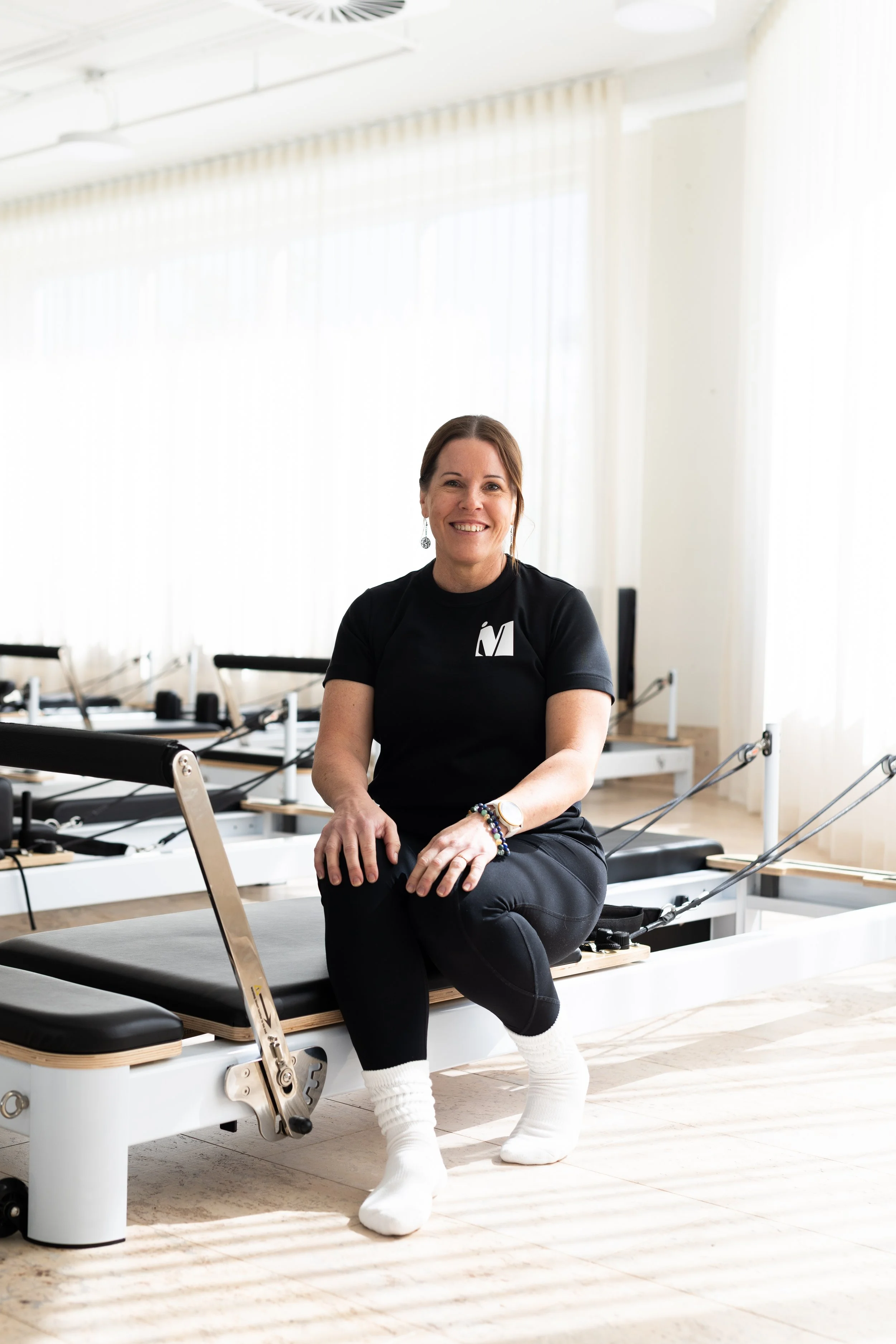A woman standing indoors beside a wooden console table, smiling, wearing a black t-shirt with white lettering, black leggings, and black shoes, with three framed photographs hanging on a white wall behind her.