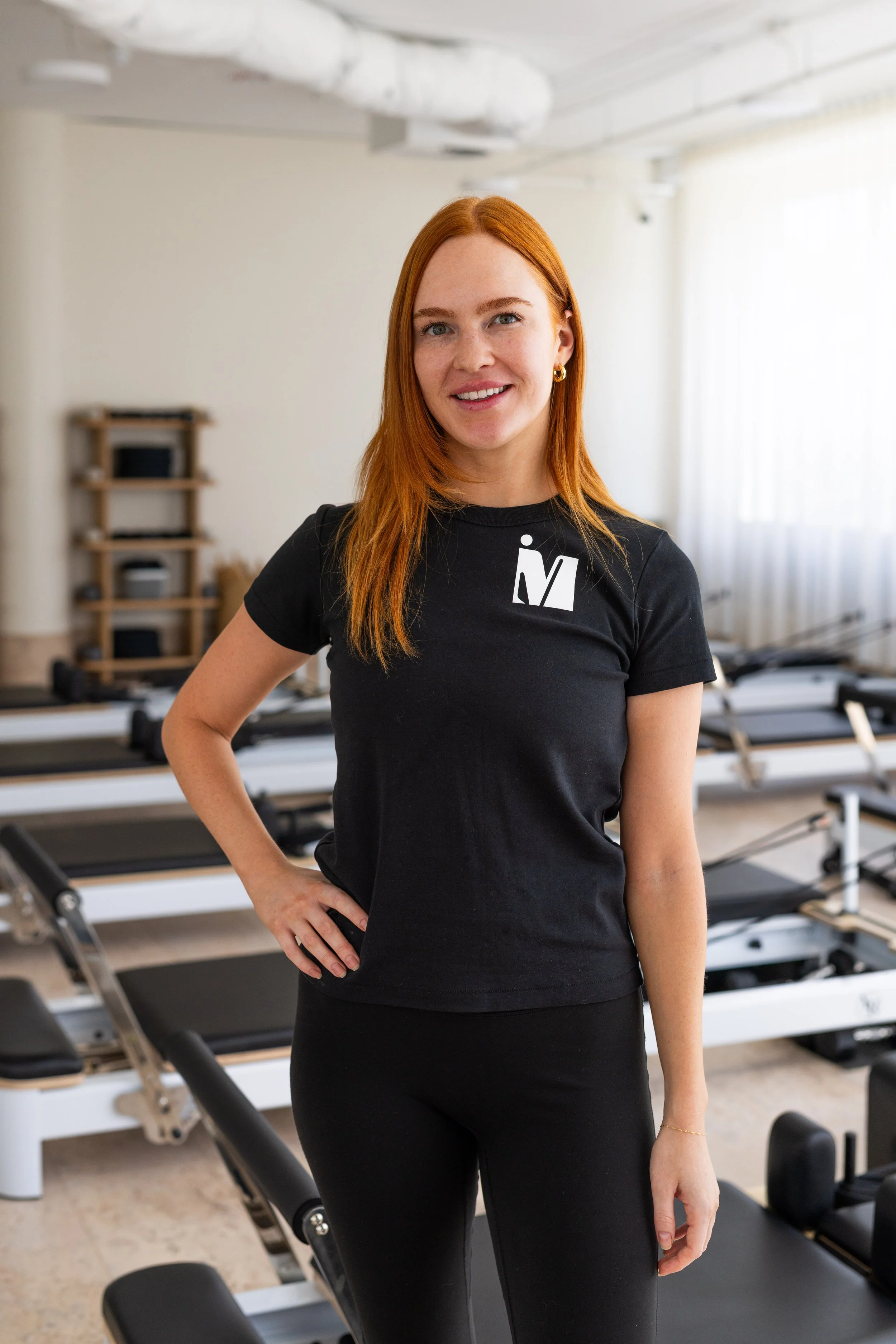 A woman with red hair sitting on a Pilates reformer machine in a fitness studio with mirrors and modern lighting.