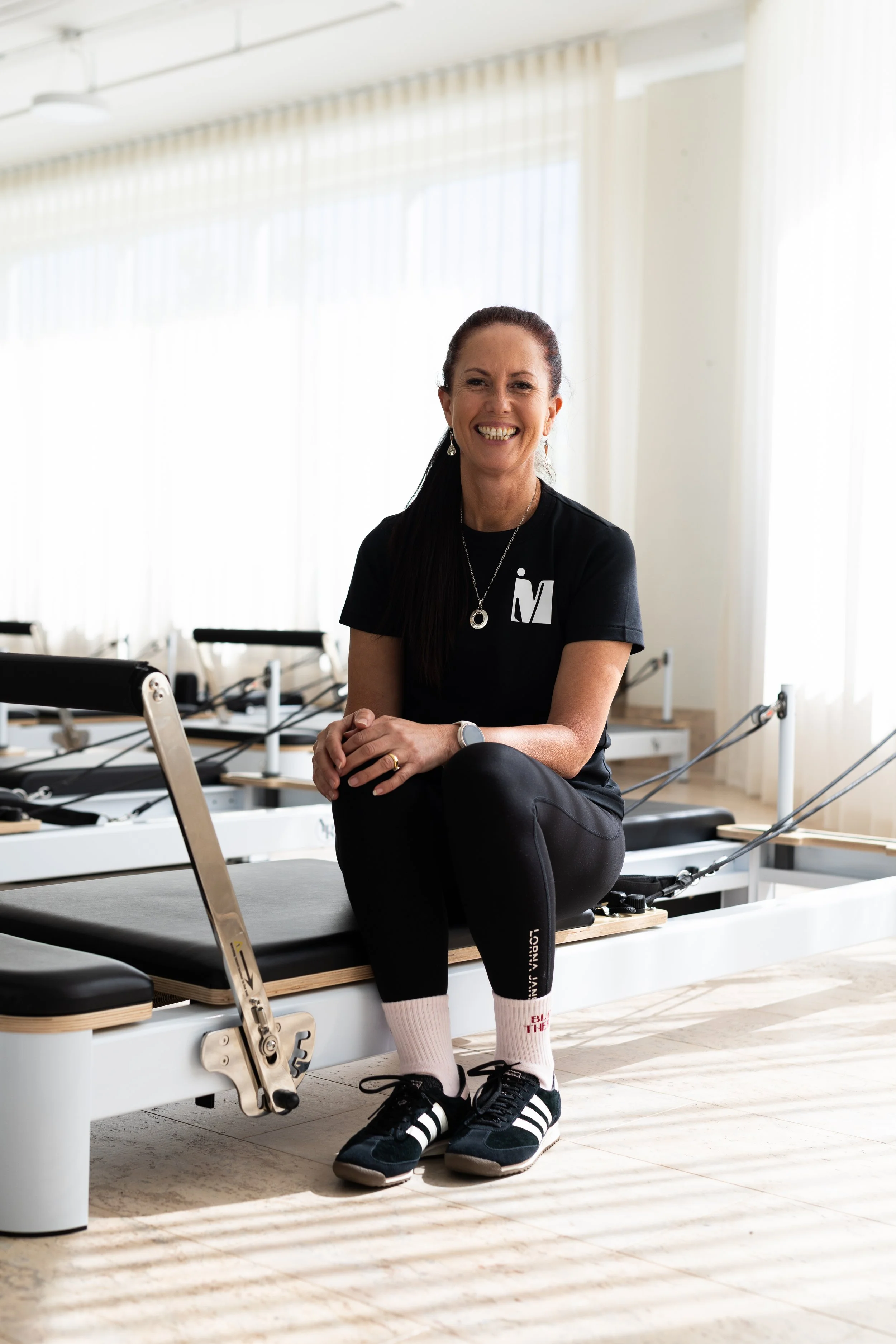A smiling woman standing in a Pilates studio with reformer machines arranged in rows behind her.