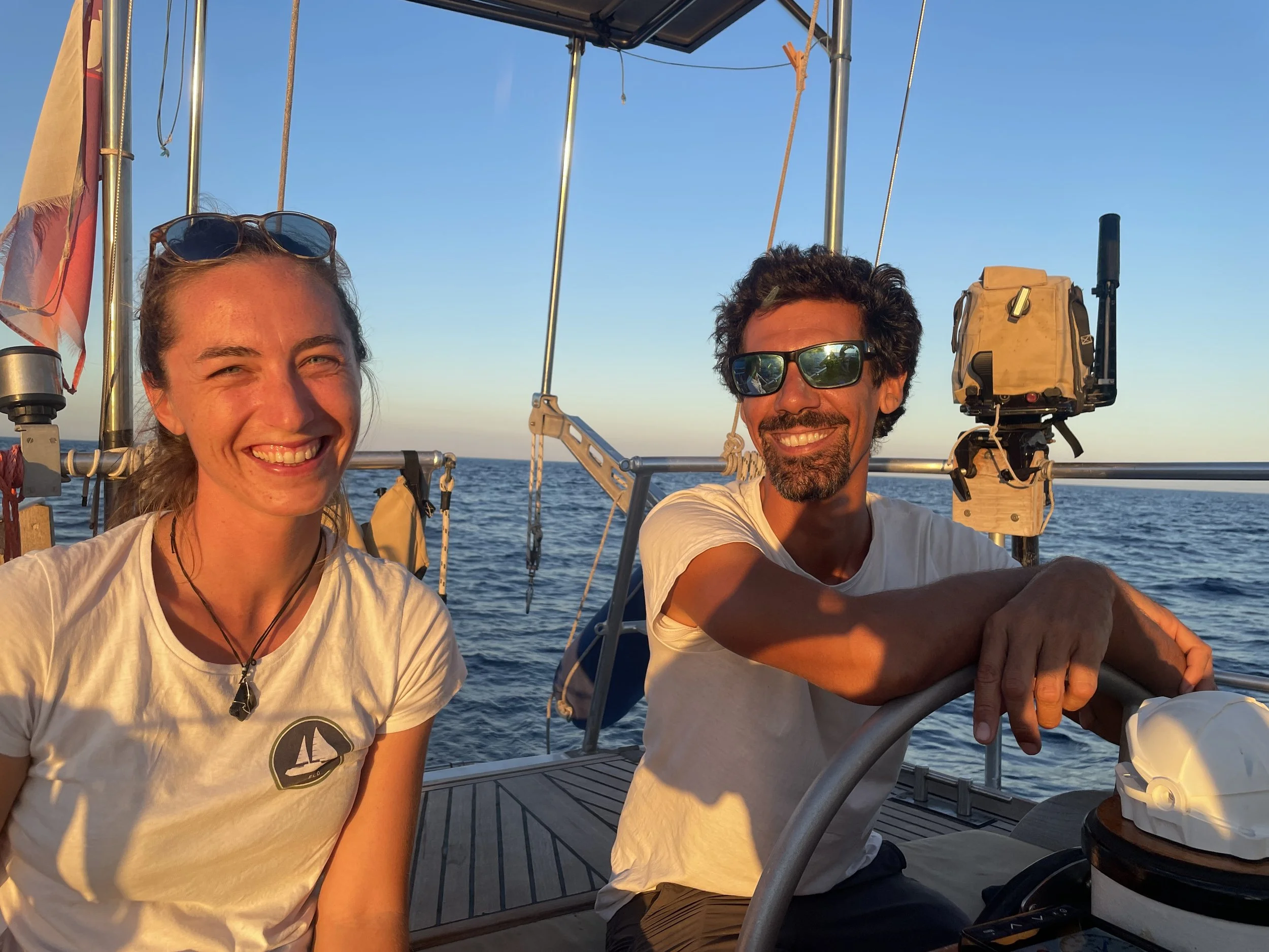 Two smiling people with sunglasses on a sailboat at sunset, with the ocean in the background.
