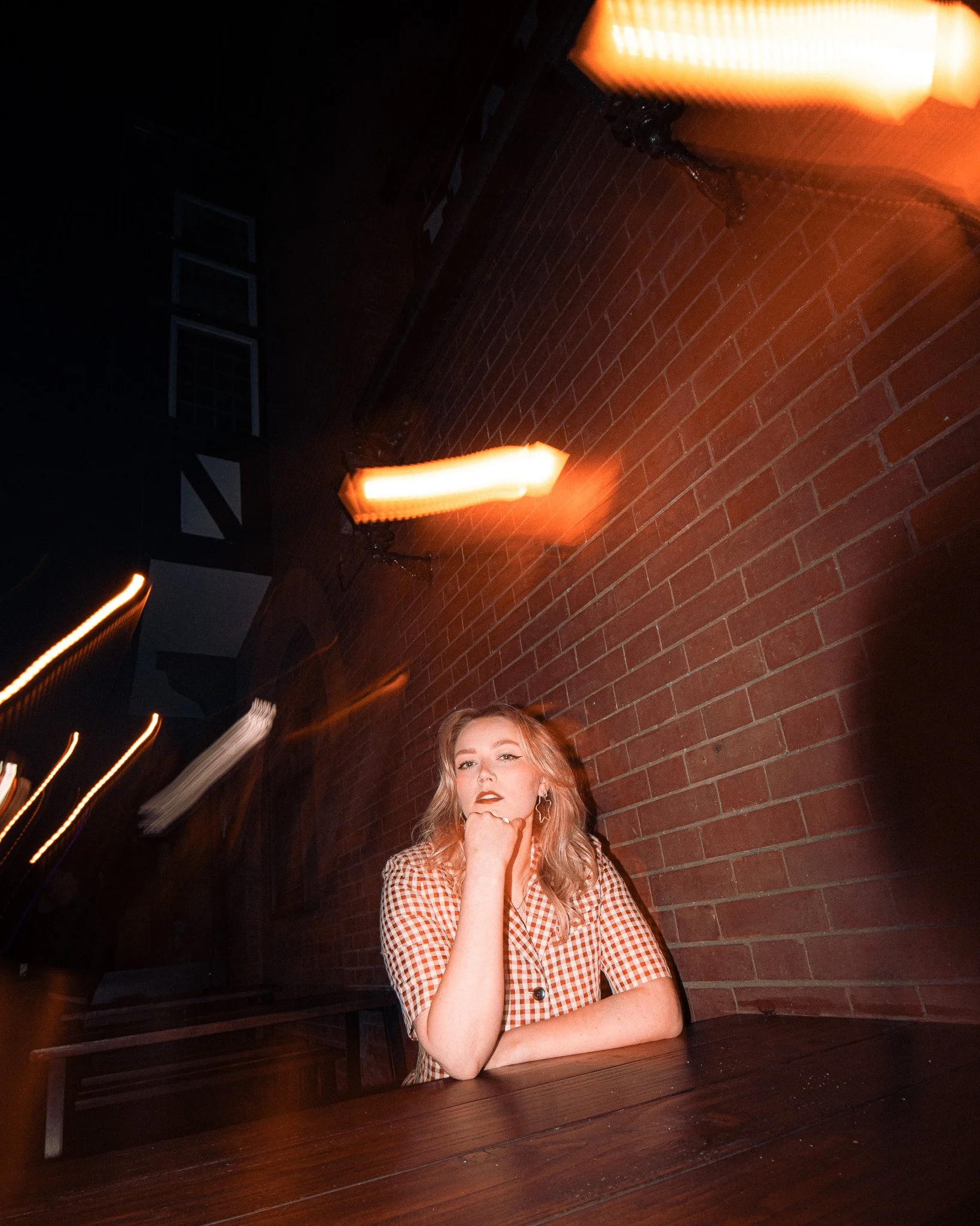 Hannah Michell before her set at the Hare & Hounds, Kings Heath, Birmingham. A young woman with blonde hair sitting at a table in a dimly lit room with a brick wall, dramatic lighting, and orange lamps.