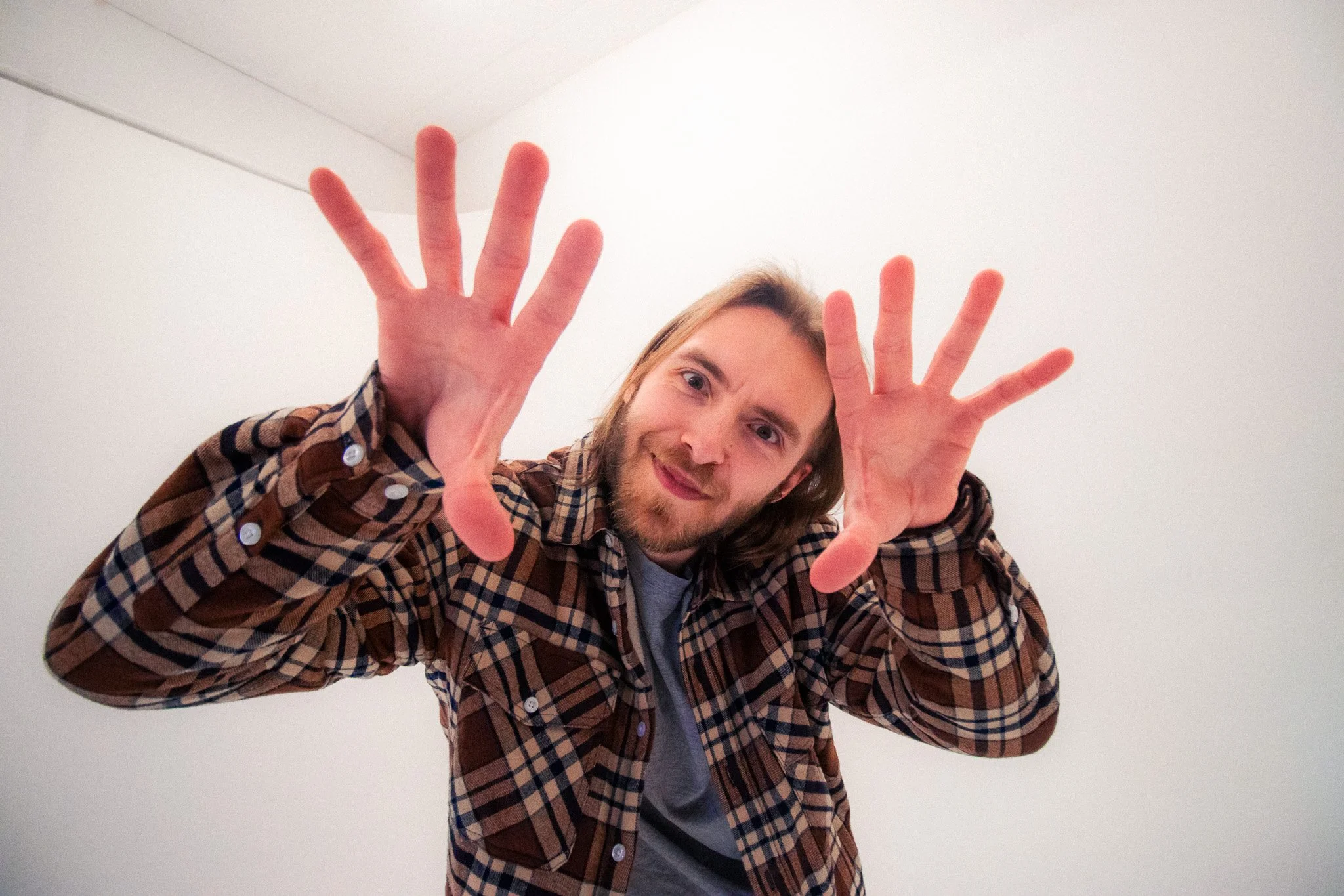 The Spoils at PHOS Studio, Digbeth, Birmingham. A young man with shoulder-length hair and a beard, wearing a checkered brown jacket over a gray shirt, is smiling and leaning towards the camera.