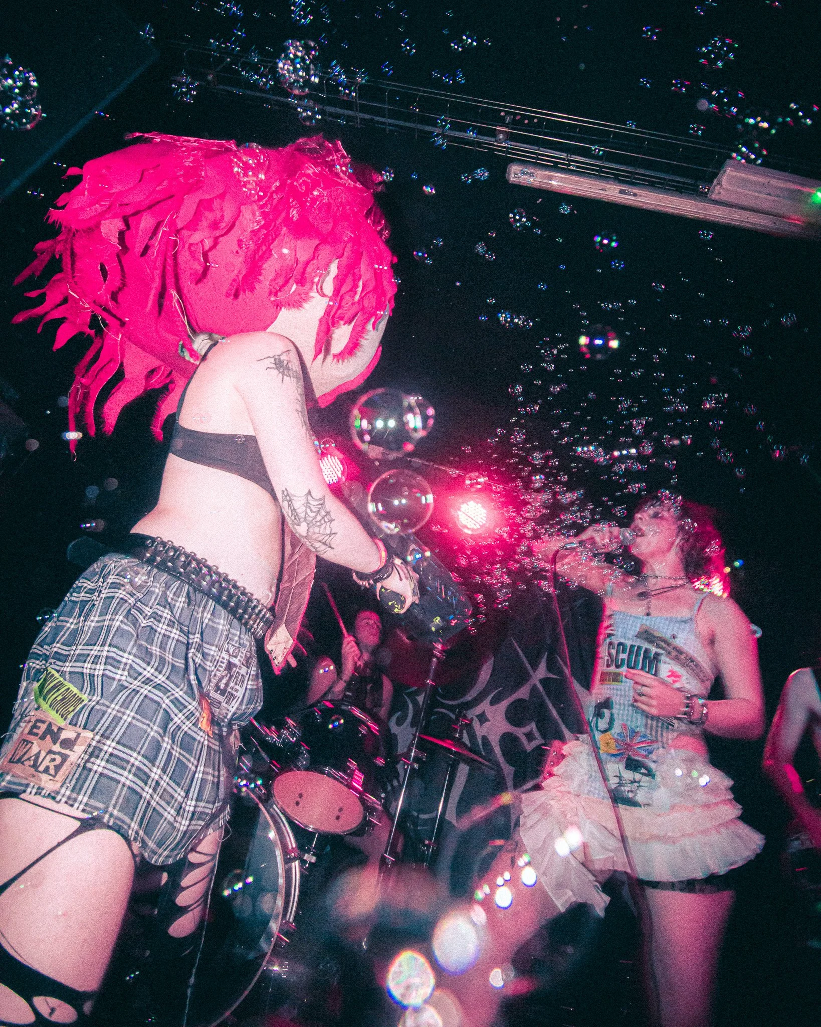 Bex at The Sunflower Lounge. A band performing on stage with pink hair, tattoos, and punk style clothing, surrounded by bubbles and pink lighting.