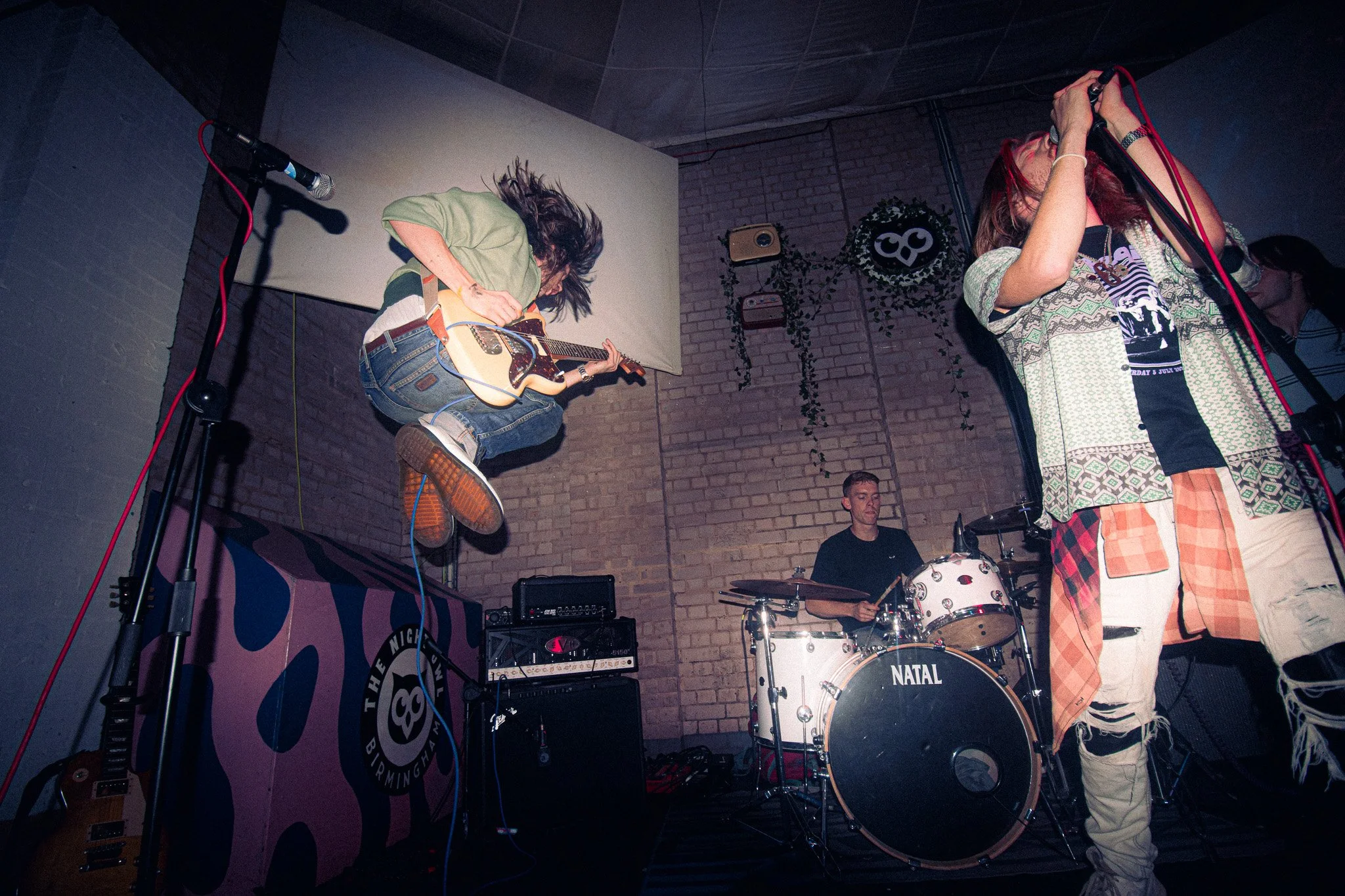 Fuel Injection, The Night Owl. Digbeth, Birmingham. 
A band performing live on stage with a guitarist jumping, a singer holding a microphone, and a drummer playing in the background, in a cozy venue with brick walls and stage equipment.