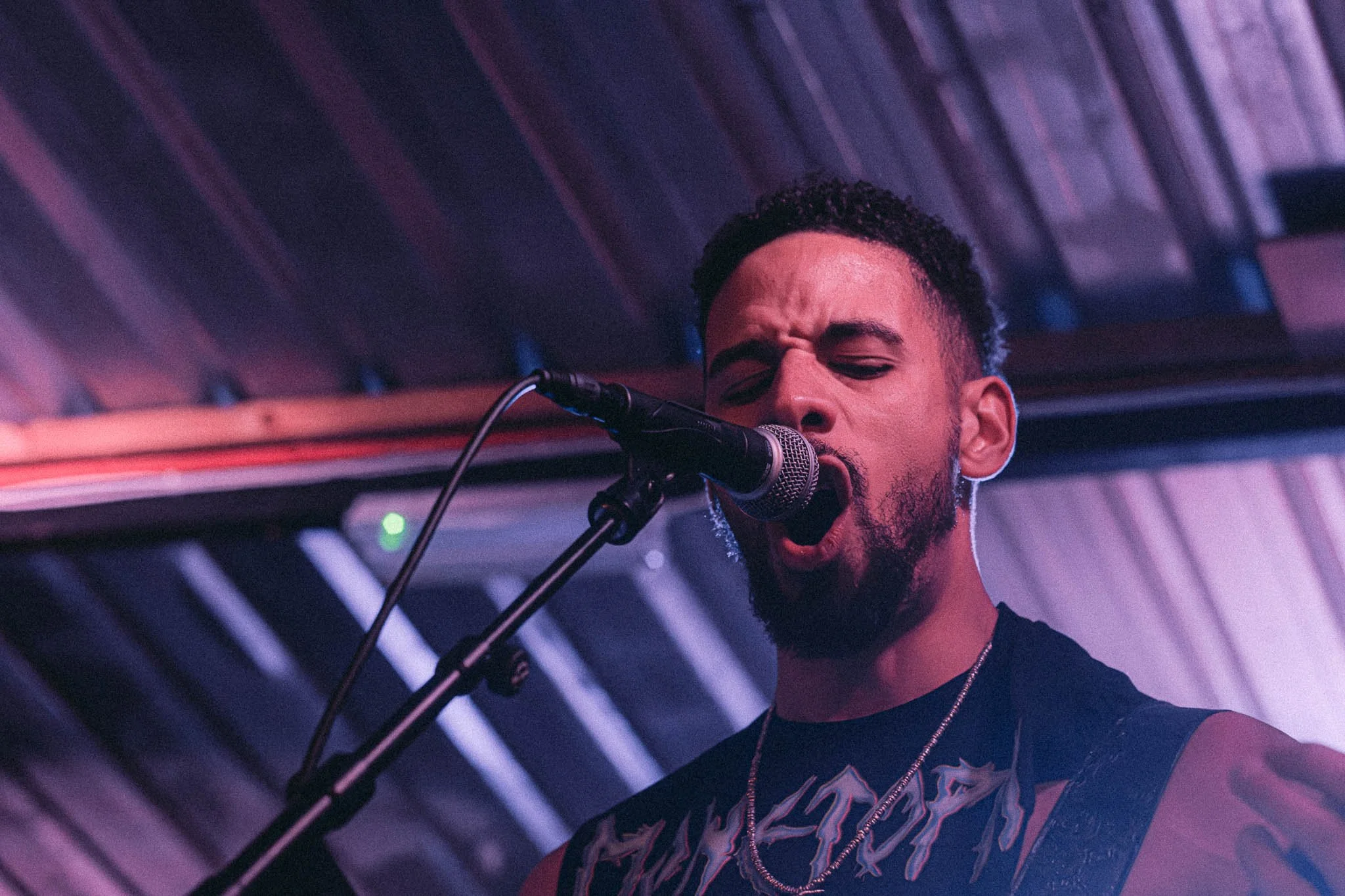 Monetopia at Dead Wax Courtyard, Digbeth, Birmingham. Male singer with a beard singing passionately into a microphone at a live music venue with wooden ceiling panels.