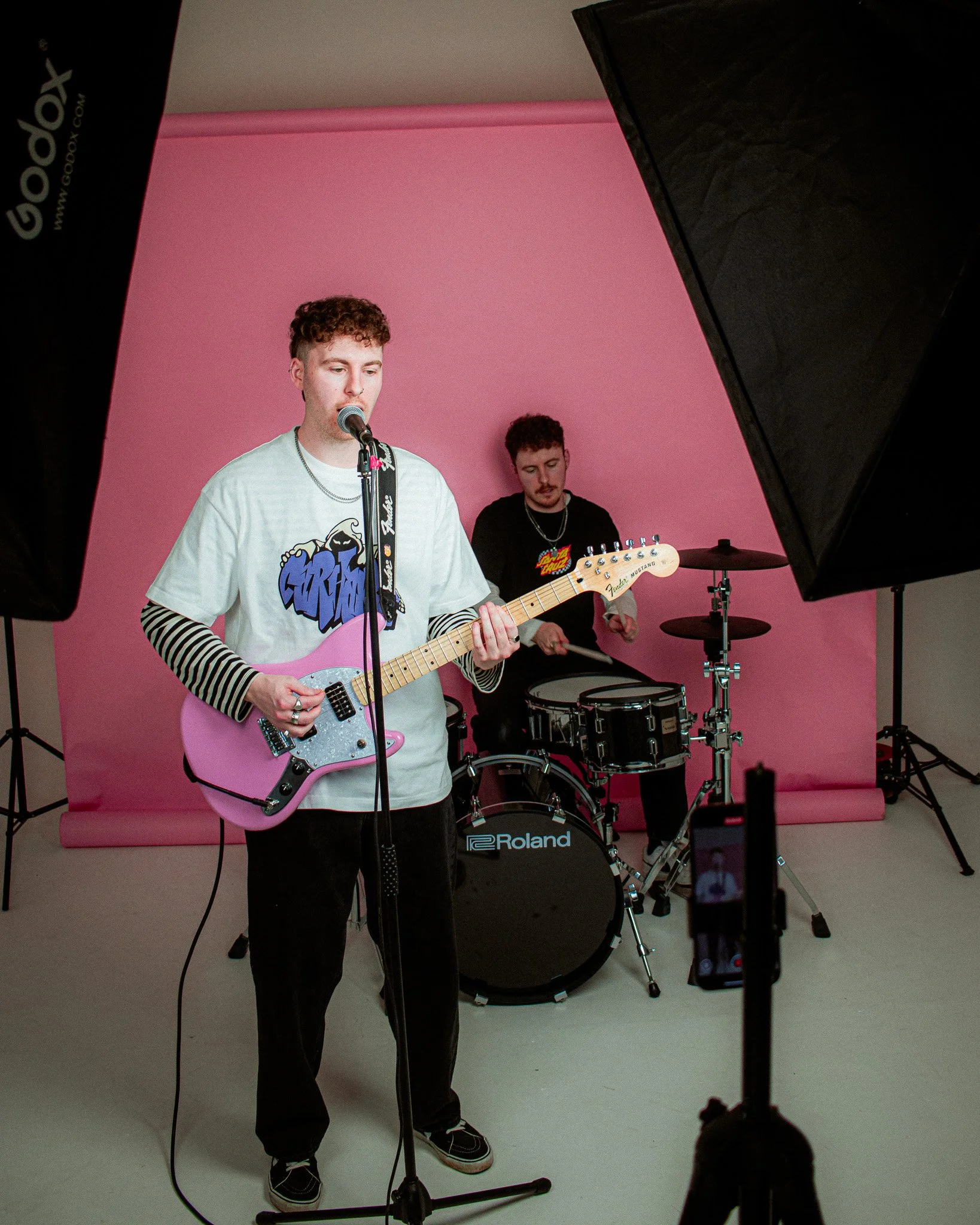 Twinninetysix at PHOS studio, Digbeth, Birmingham. Two young men performing music in a studio with a pink background. One is singing and playing a pink electric guitar, the other is playing drums.