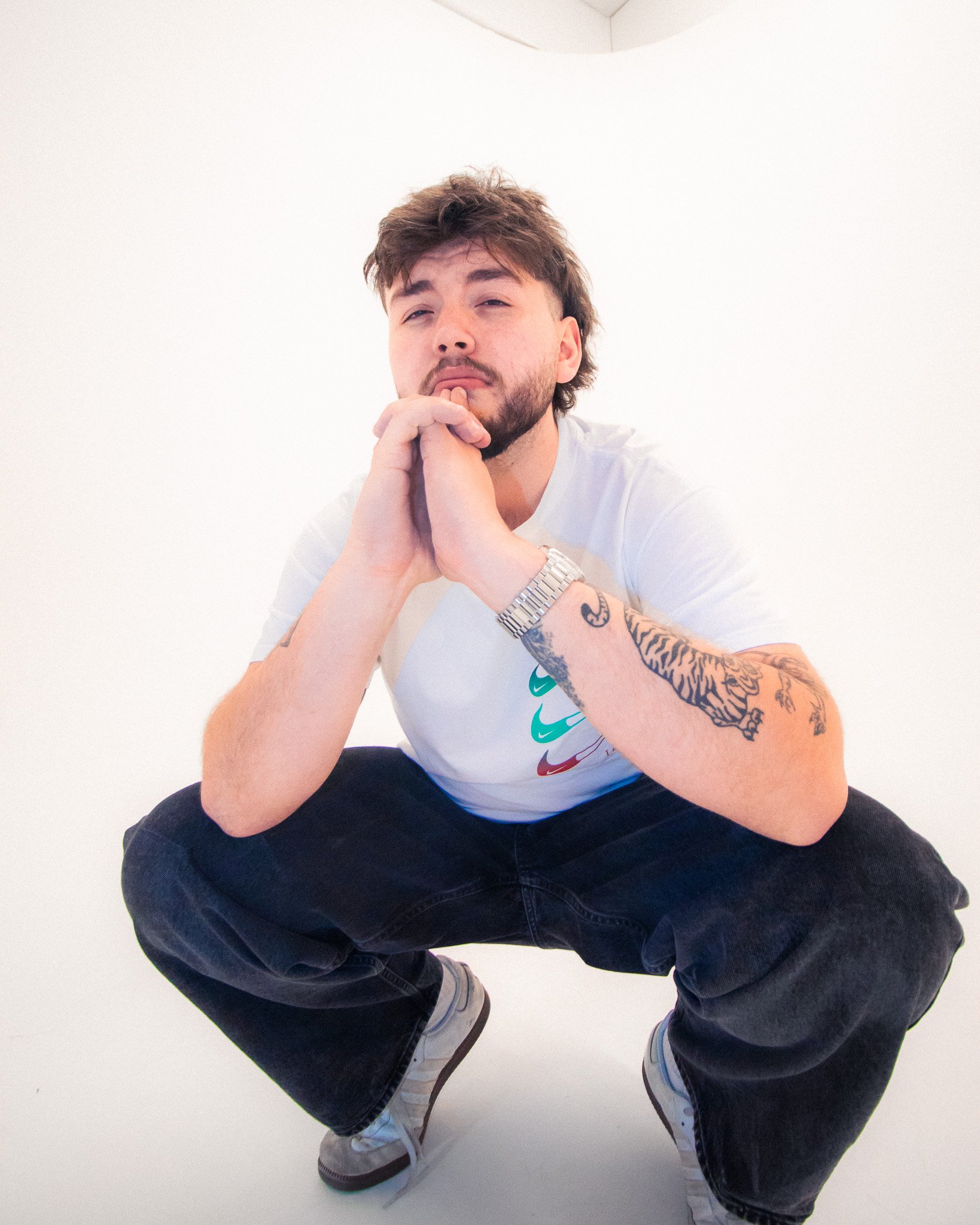 The Spoils at PHOS Studios, Digbeth, Birmingham. Young man squatting in front of a plain white background, wearing a white t-shirt, dark jeans, sneakers, and a wristwatch, with beard, tattoos, and styled hair.
