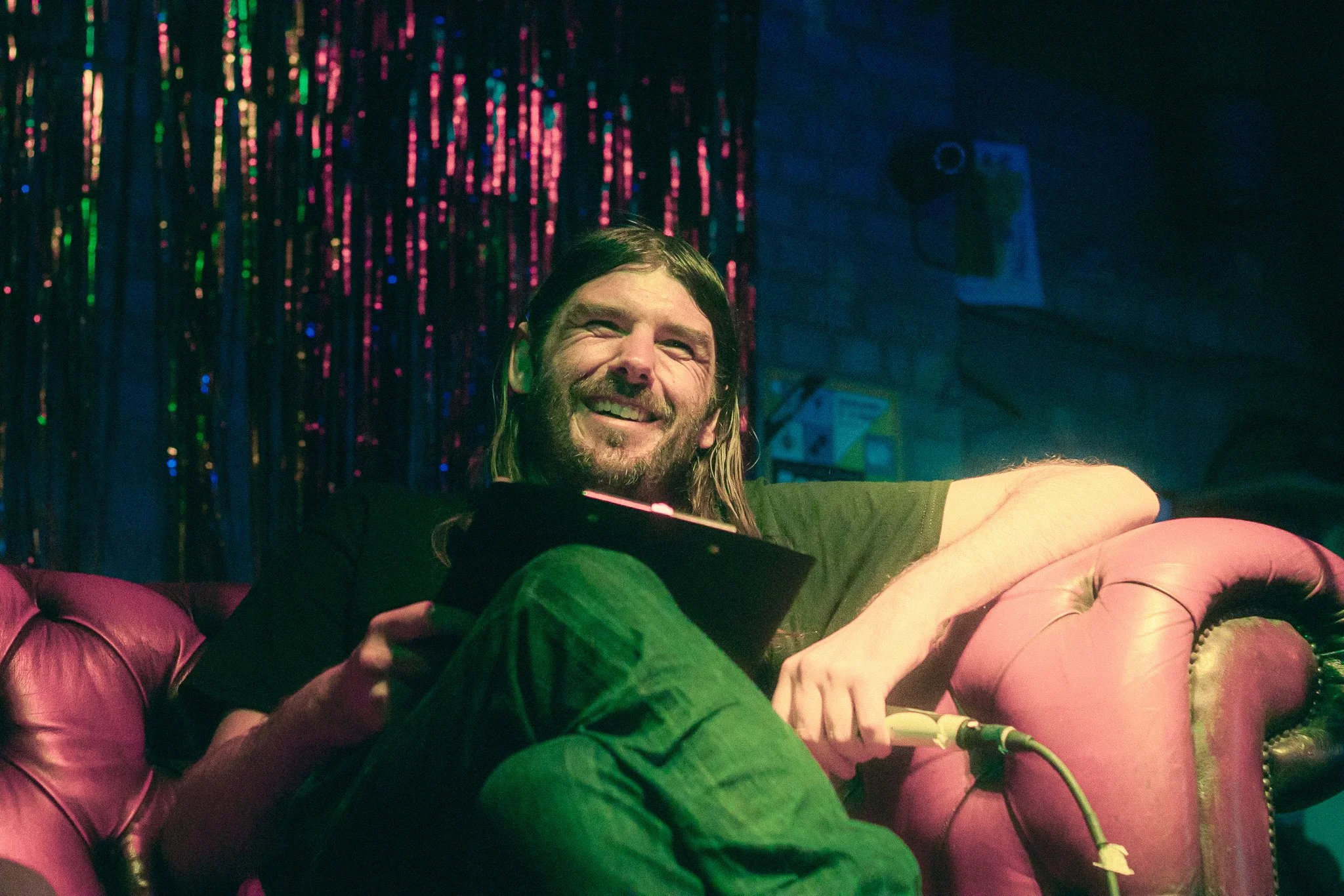 DJ Millabong during Let's Build an Alternative Network at The Night Owl, Digbeth, Birmingham. A man with long hair and a beard is sitting on a pink leather couch, smiling, and holding a clipboard.