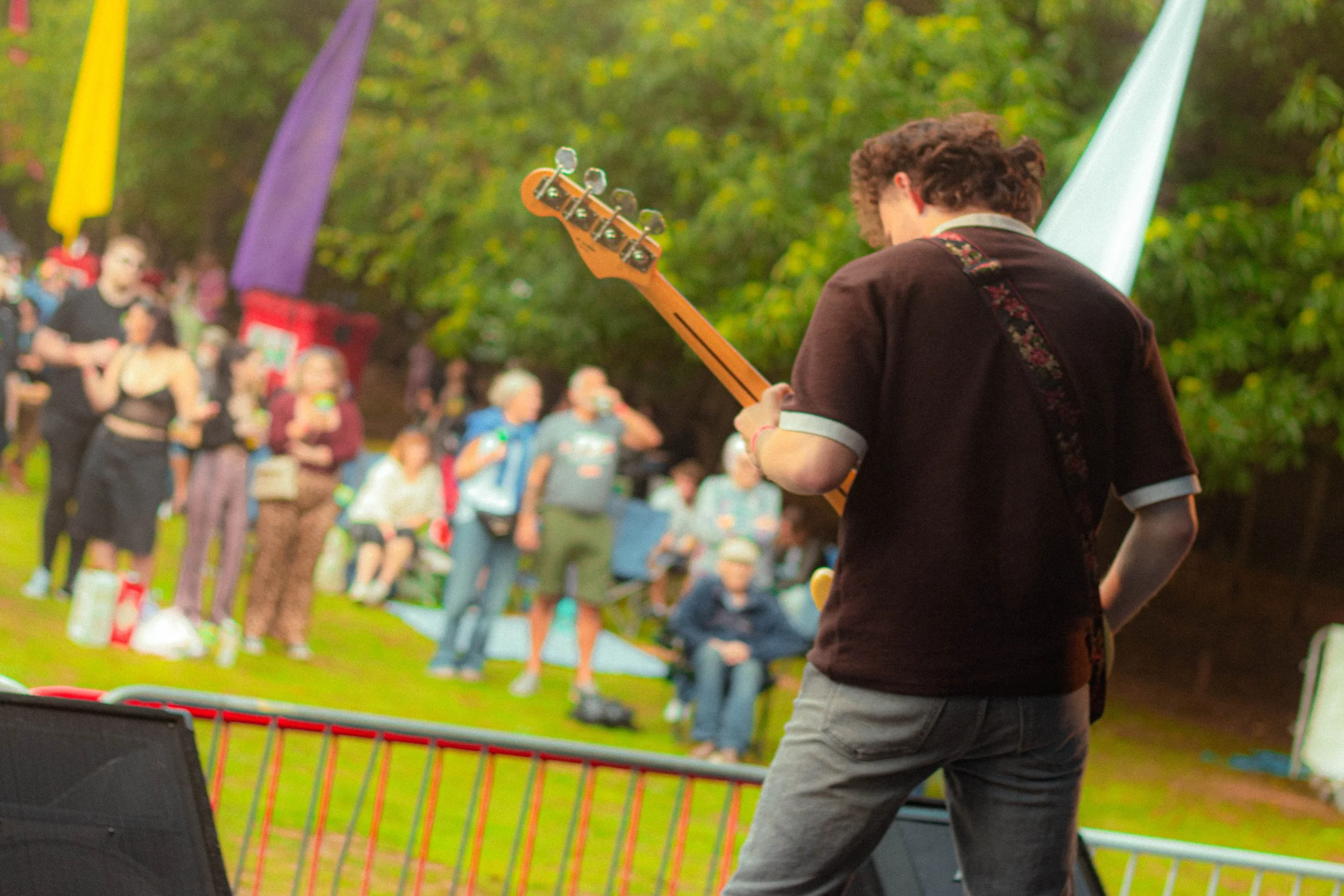The Spoils in Codsall, performing at Codfest. Musician playing electric guitar on stage, audience watching at outdoor event with trees and colorful flags in background.