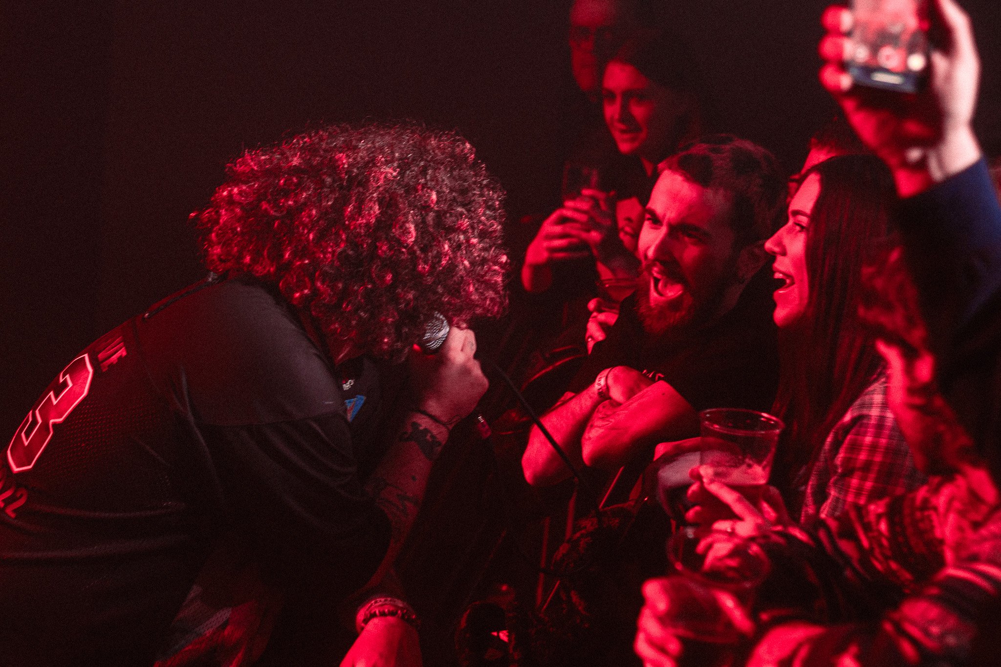 Santalum at o2 Institute3, Birmingham. A performer with curly hair and tattoos on their arm performs with a microphone for a crowd. The audience appears excited, with one person holding a drink and others cheering or smiling.