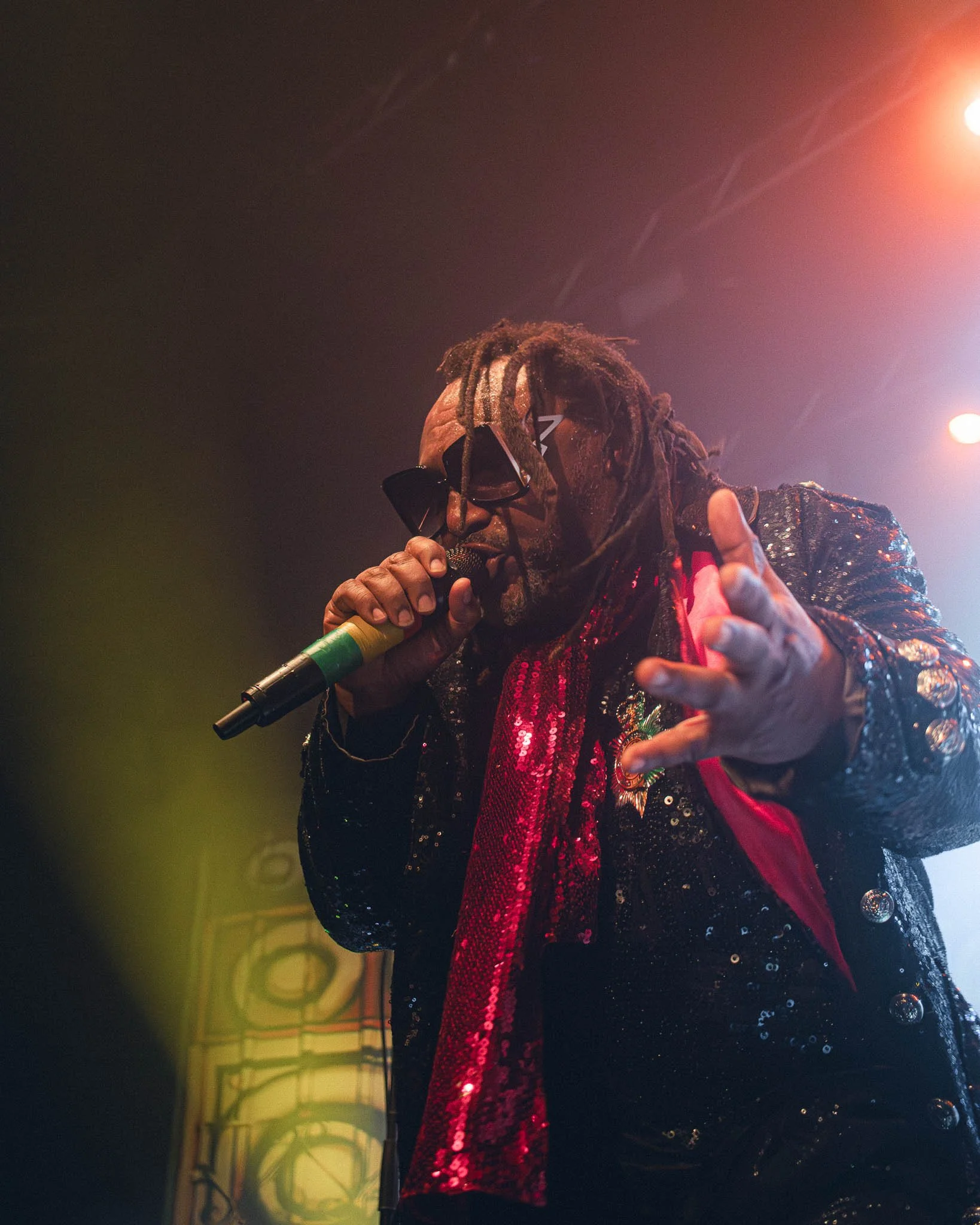 Skindred at KK's Steel Mill, Wolverhampton. A performer with dreadlocks, wearing sunglasses and a sequined outfit, singing into a microphone on stage with colorful lighting.