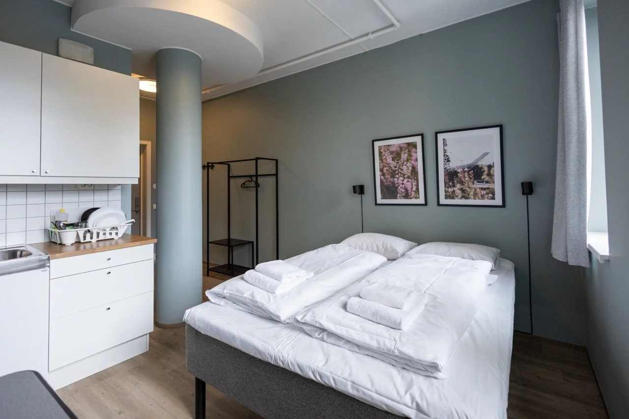 Practical private room with double bed and a small kitchenette area with plates and dishes, and an open clothing rack in the background. Affordable hotel in Oslo