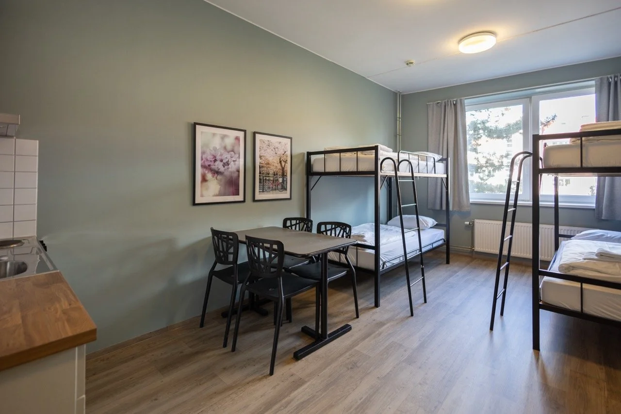 Practical private room with four bunk beds, a small dining table with four black chairs, light green walls, a large window with curtains, and framed artwork on the wall. Private hostel room in Oslo