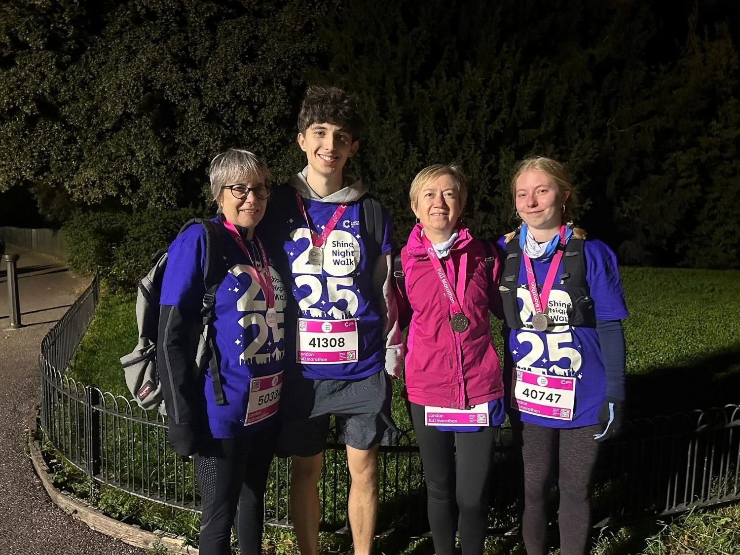 Four people wearing purple Shine Night Walk 25 shirts and finisher medals stand together in a park at night after completing the London Shine Walk, smiling and posing for a photo.