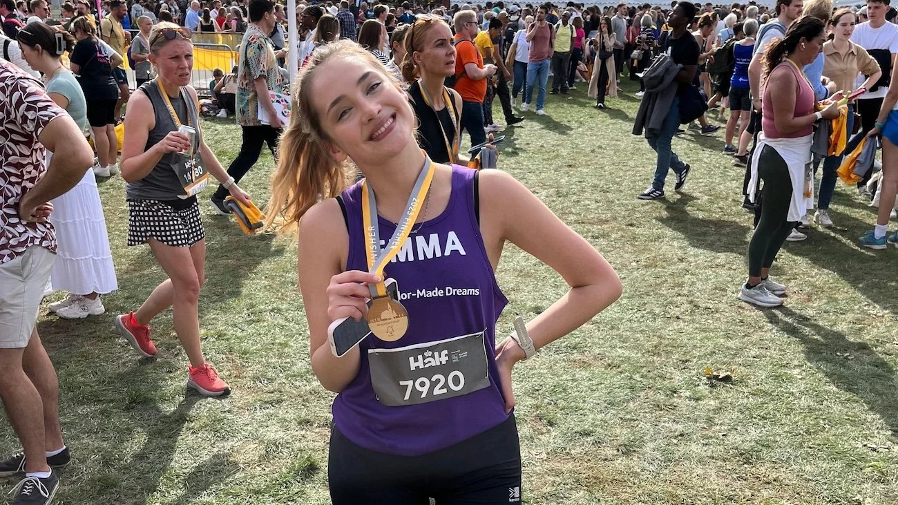 A smiling runner wearing a purple charity vest holds up a finisher medal after completing a half marathon, standing in a busy finish area surrounded by other participants.
