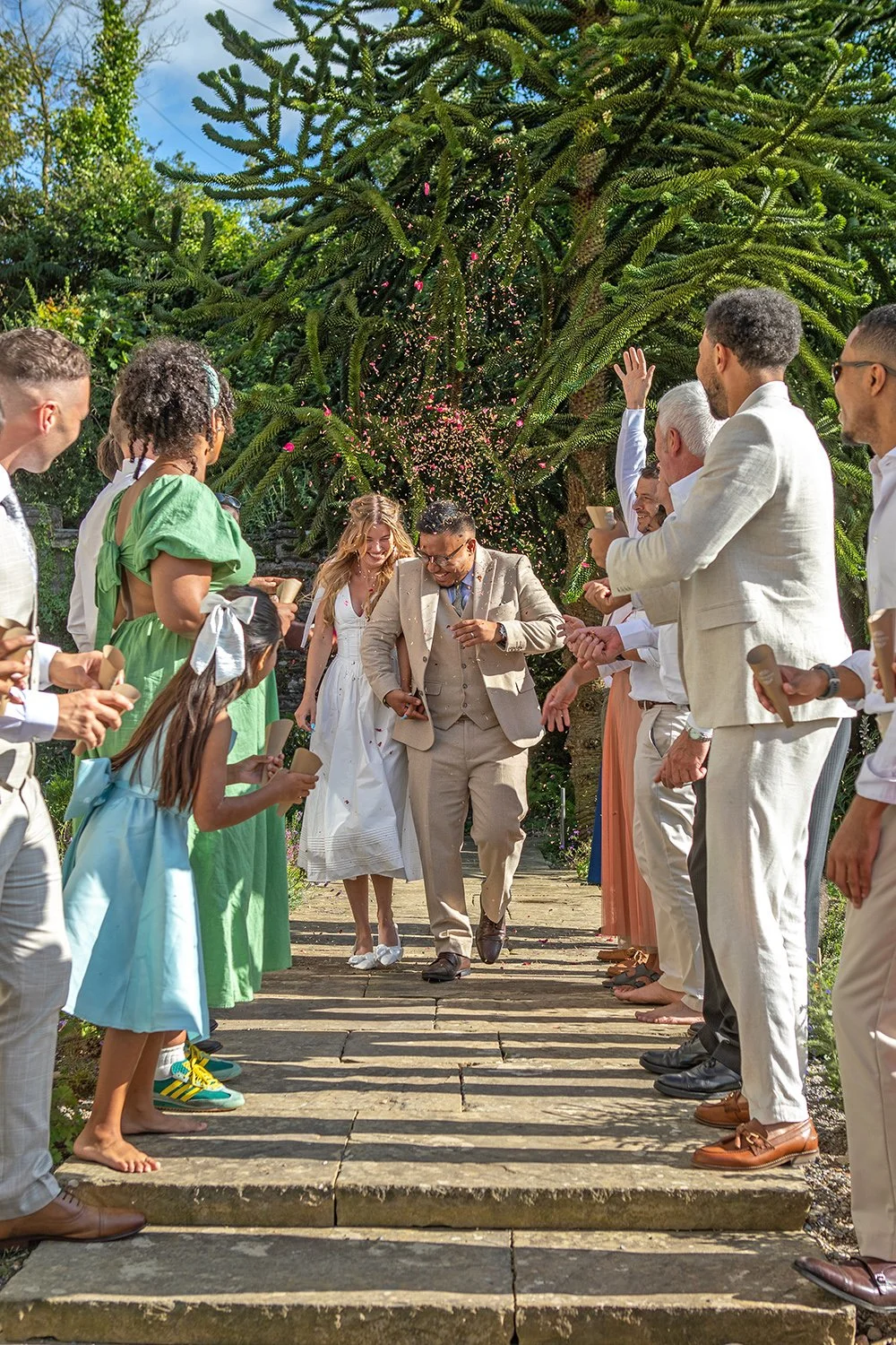 A diverse group of people celebrating a wedding outdoors on a sunny day. They are dressed in semi-formal and casual attire, smiling and holding confetti and party poppers as they walk down a stone pathway.