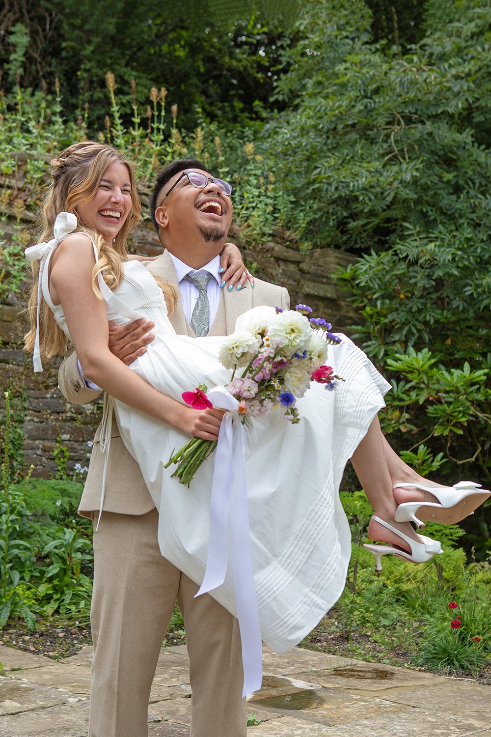 A bride and groom, the bride carrying a bouquet of flowers, outdoors with greenery in the background. Both are smiling and laughing, celebrating a joyful event.