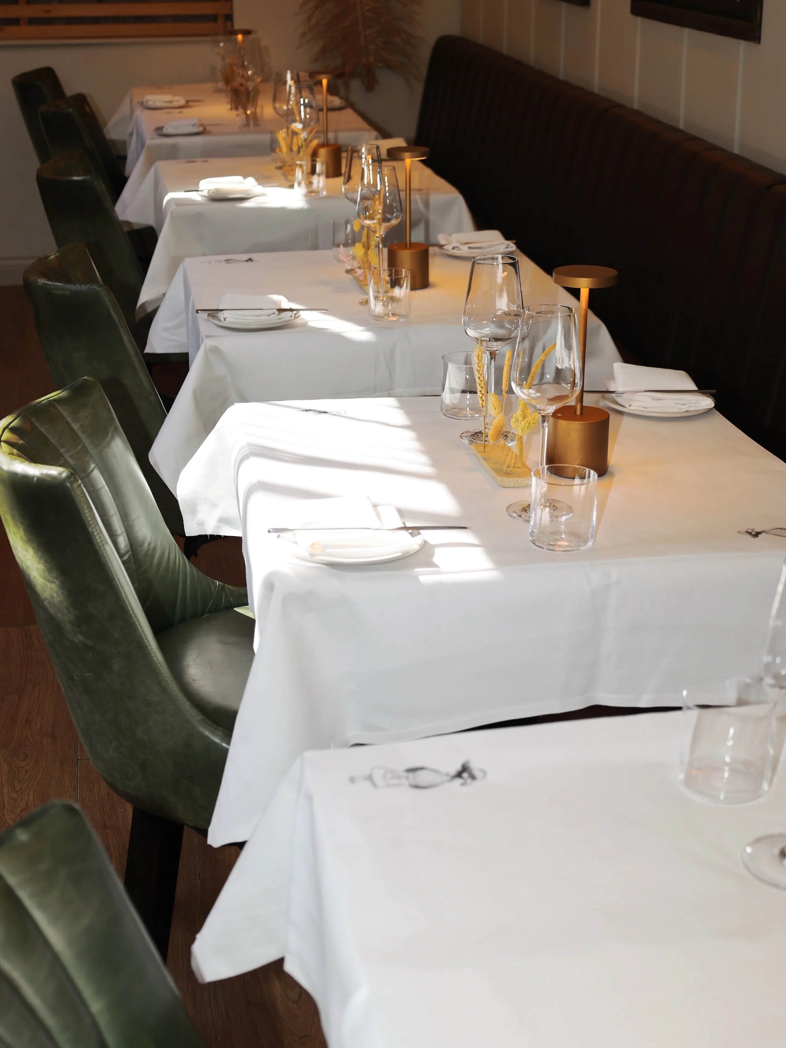 Empty restaurant tables with white tablecloths, set with plates, glasses, and silverware, illuminated by natural light.