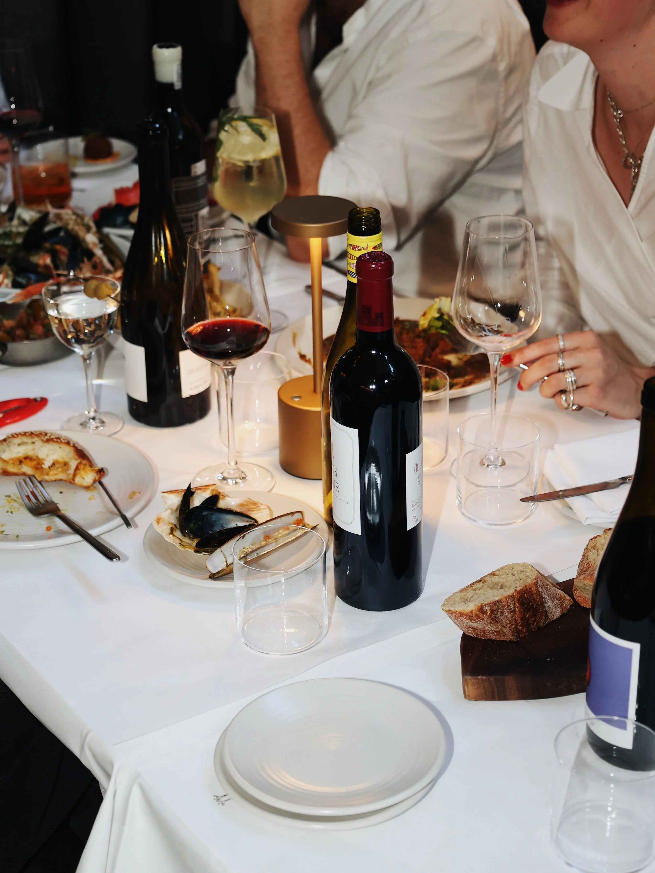 A dining table with wine bottles, glasses of red and white wine, a bottle of yellow condiment, a loaf of bread, and partially eaten plates of food, with people dressed in white shirts seated around.