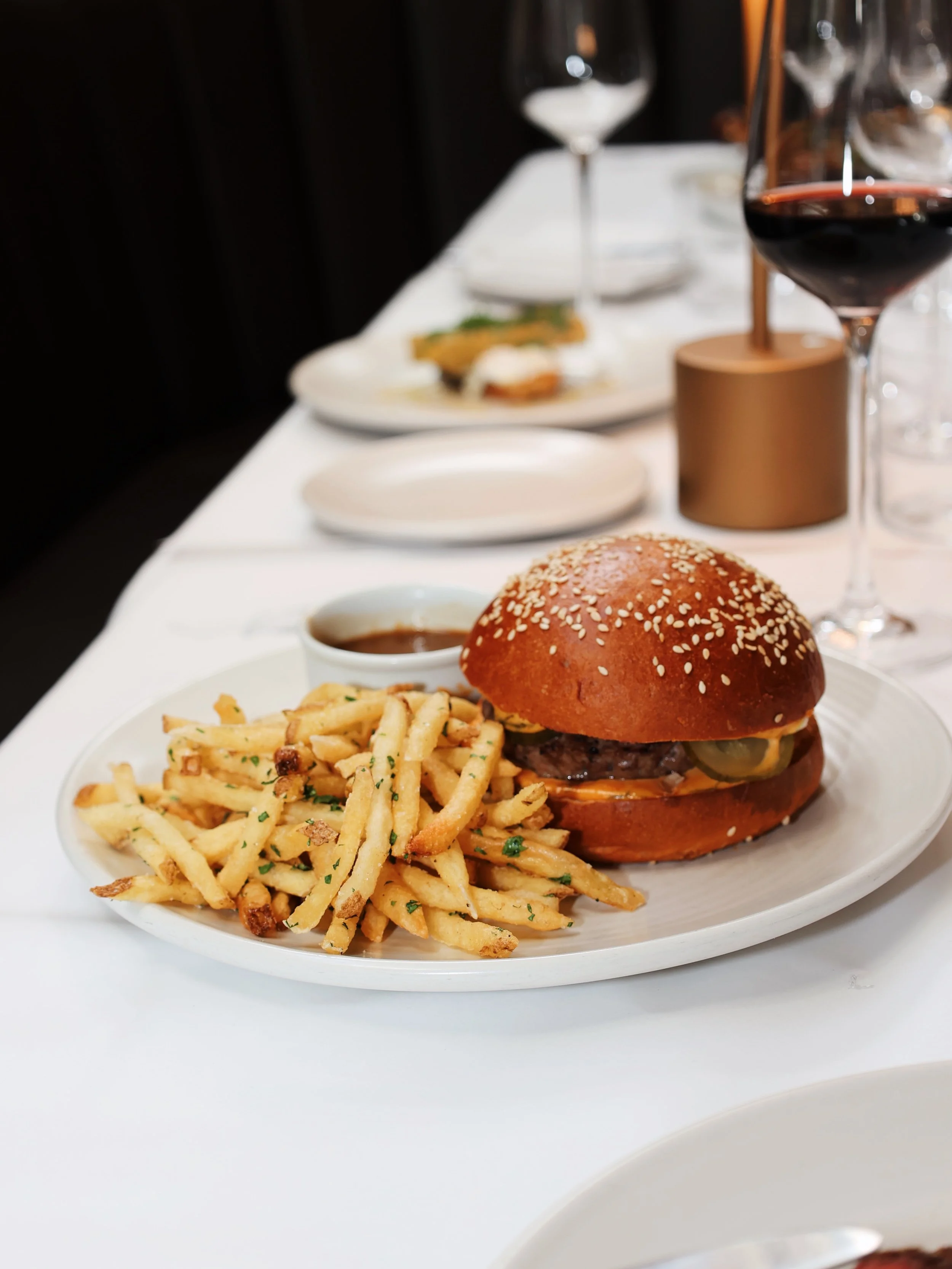 A plate of French fries and a cheeseburger with pickles and sauce, served with a small cup of dipping sauce, on a white table in a restaurant setting. In the background, there are glasses of red wine and water, and dishes with food.