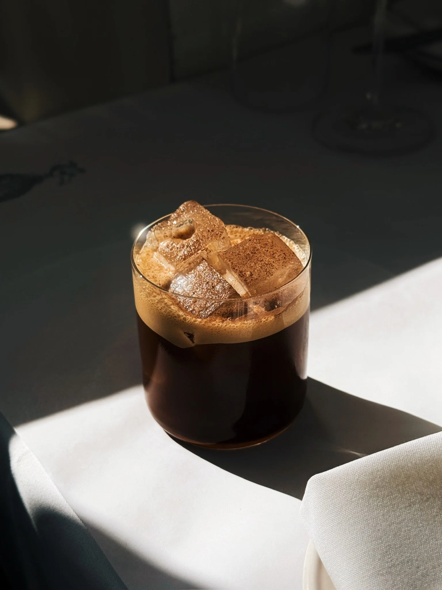 A glass of dark iced coffee with ice cubes sitting on a white table, with sunlight casting shadows.