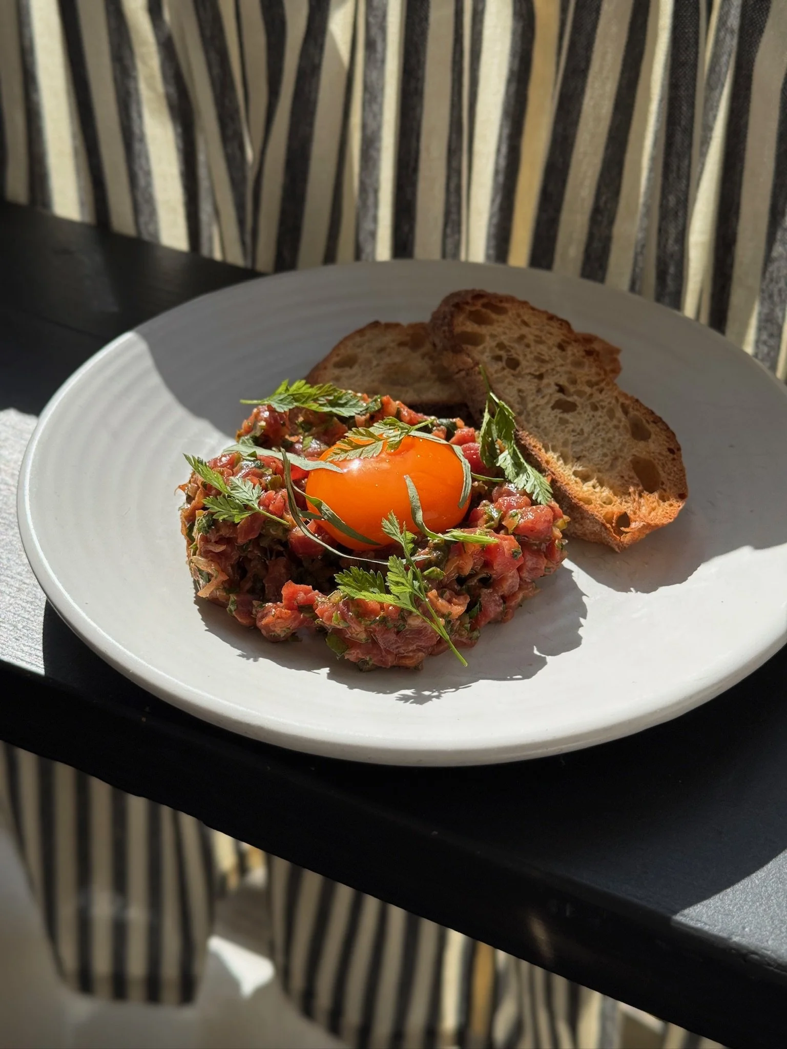 A plate of steak tartare topped with a raw egg yolk, garnished with fresh herbs, accompanied by two slices of toasted bread.