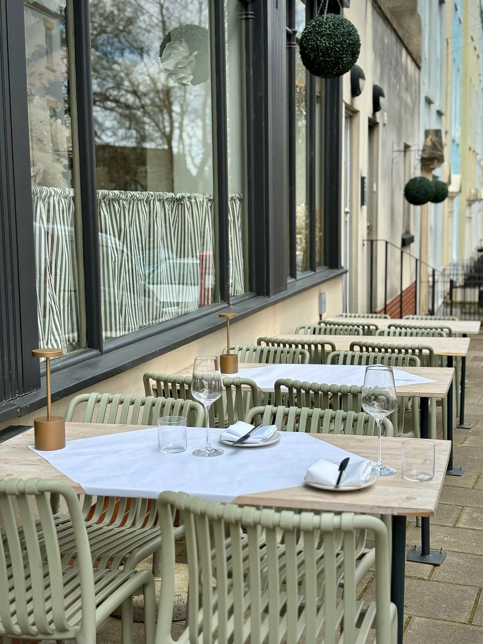 Empty outdoor restaurant tables with white tablecloths, wine glasses, and cutlery, situated on a sidewalk beside a building with large windows and hanging potted plants.