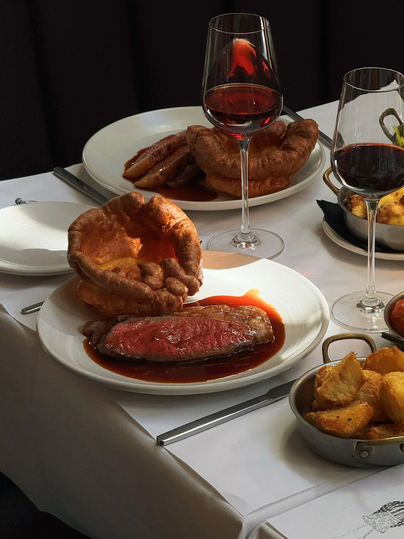 A fine dining table with dishes including roast beef with gravy in a white bowl, a Yorkshire pudding, roasted vegetables, and glasses of red wine.