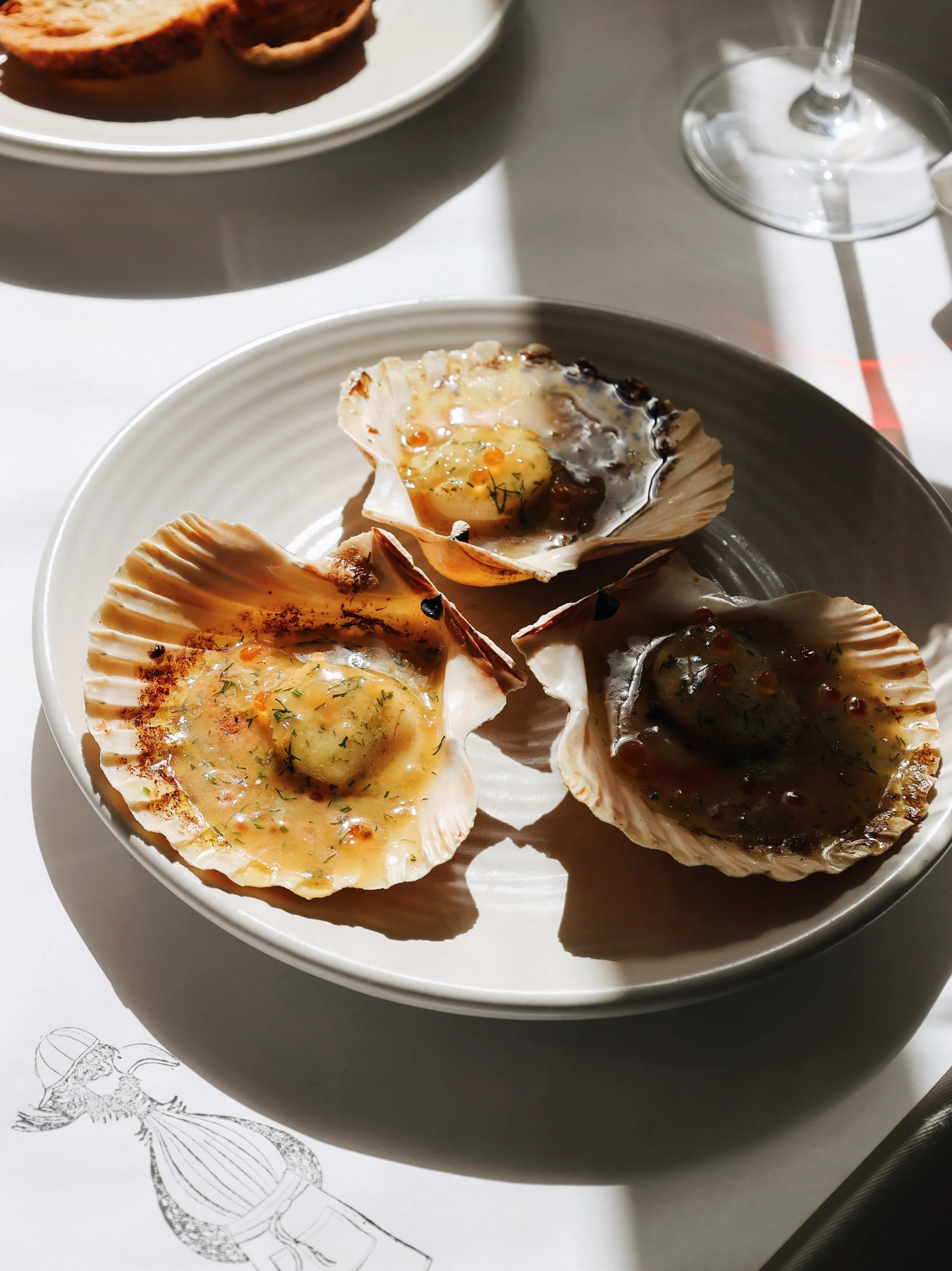 Three baked oysters with herbs and sauce on a white plate, with a glass of white wine and a partially visible plate of toasted bread in the background.