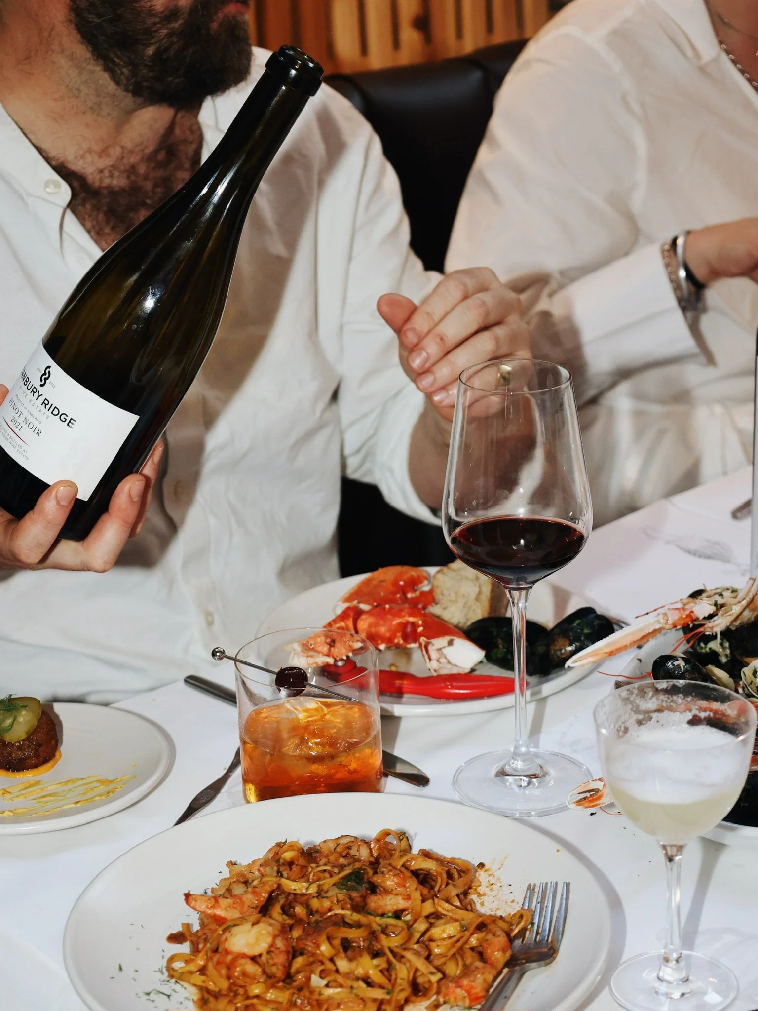 A person holding a bottle of wine above a table with various dishes, including pasta, seafood, and drinks, at a restaurant.