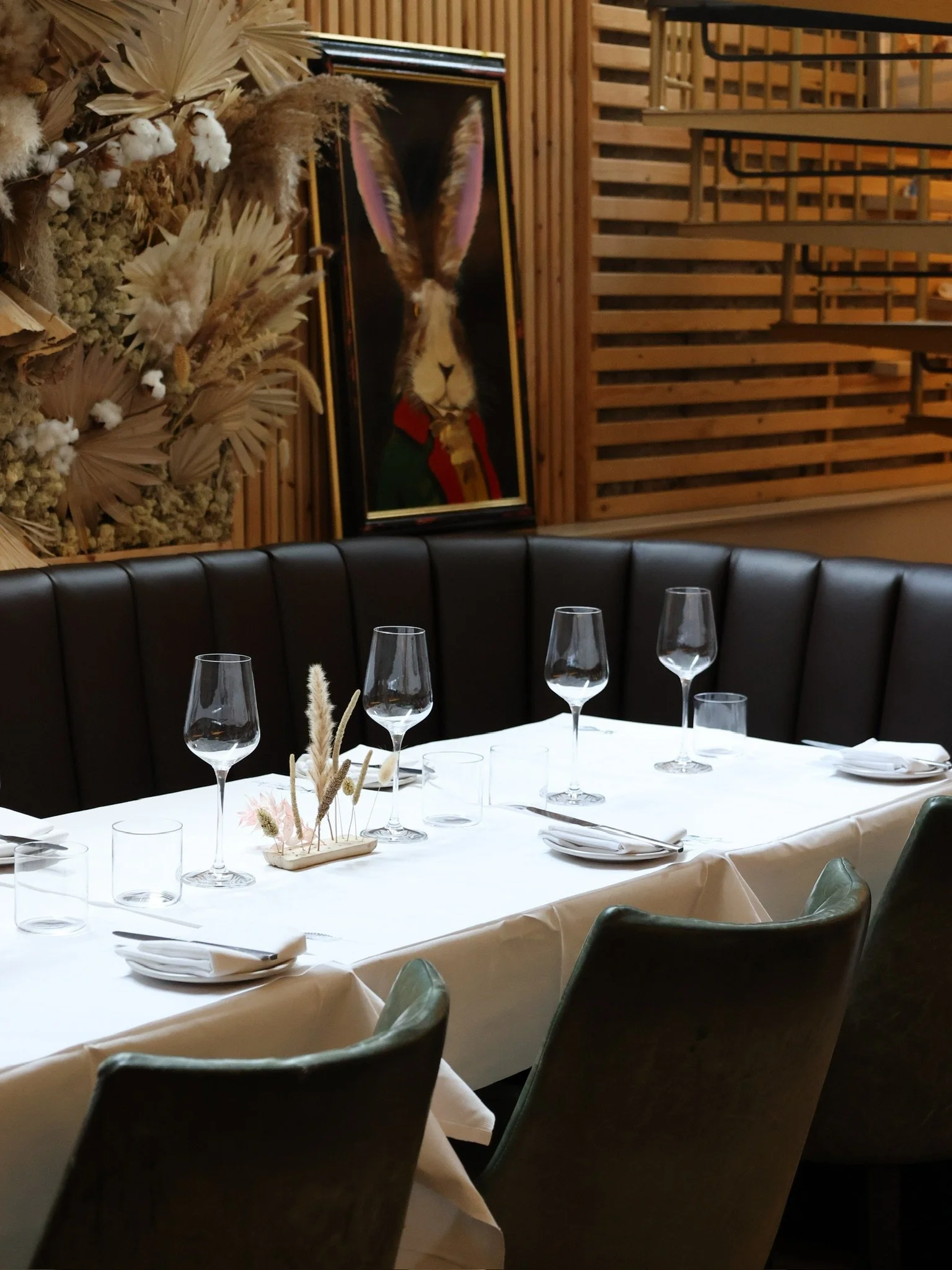 A dining table set with white tablecloth, glassware, cutlery, and a centerpiece with dried flowers in a restaurant with wooden decor and a framed painting of a rabbit with large ears wearing a red scarf in the background.