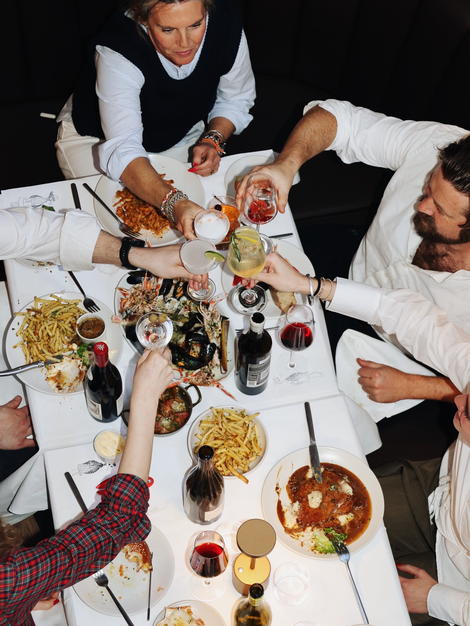 People raising their glasses for a toast at a festive dinner table with pasta, seafood, wine, and desserts.