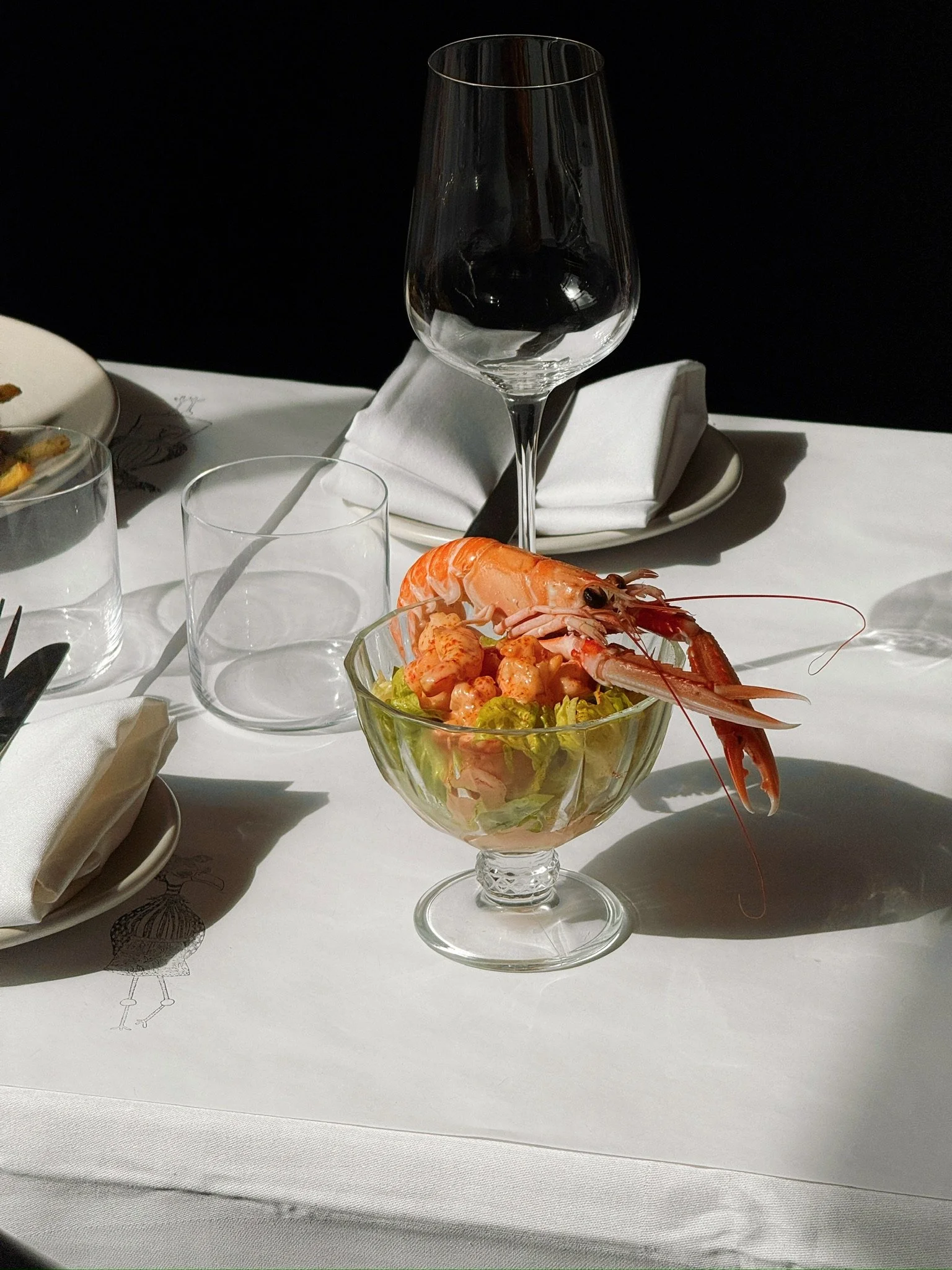 A glass bowl of shrimp salad with lettuce, topped with a whole shrimp, on a white tablecloth in a dining setting.