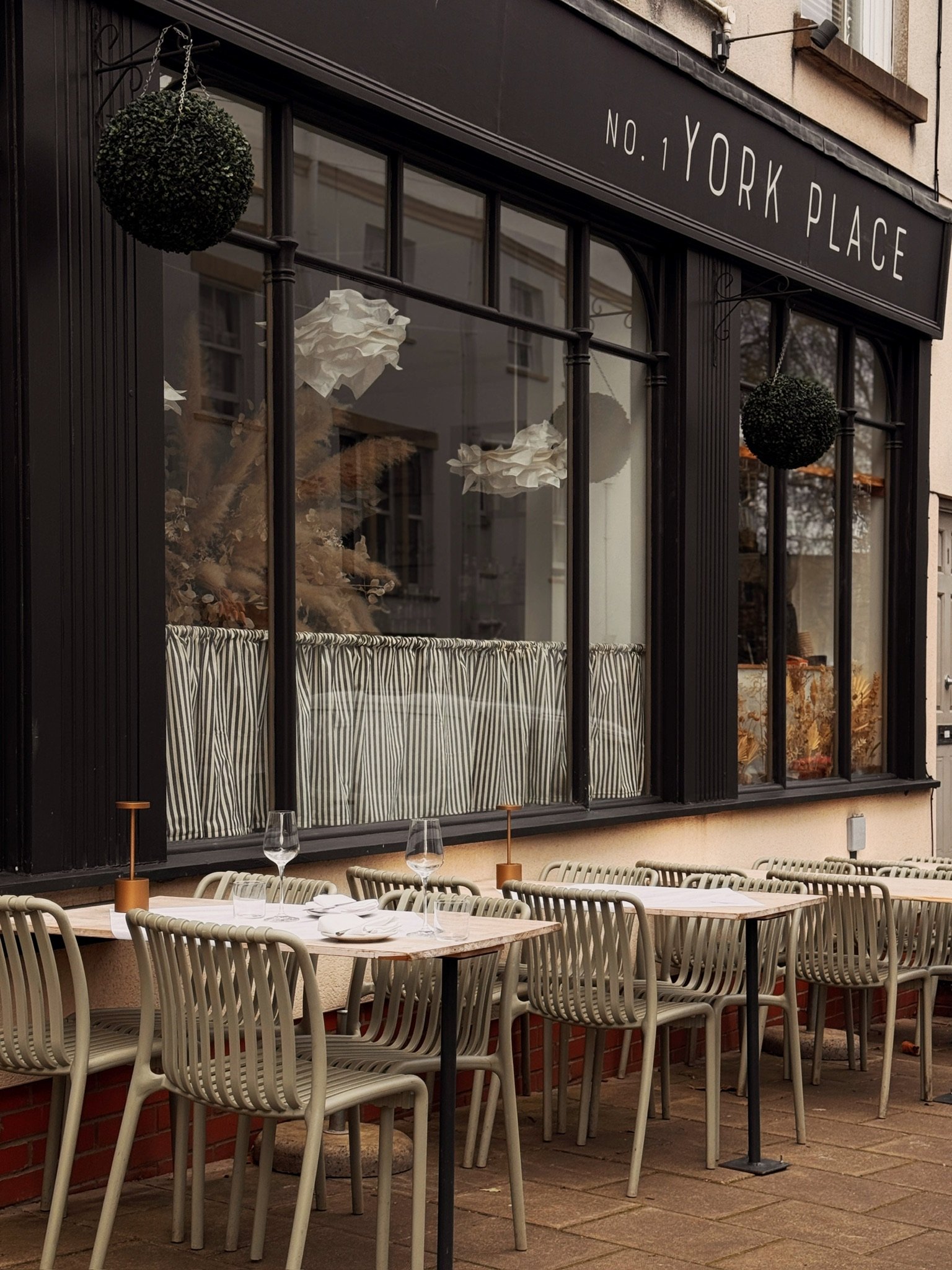 Outdoor patio dining area with tables set with white napkins, glasses, and wine glasses. The restaurant has a large black-framed window with striped curtains and hanging decorative plants.