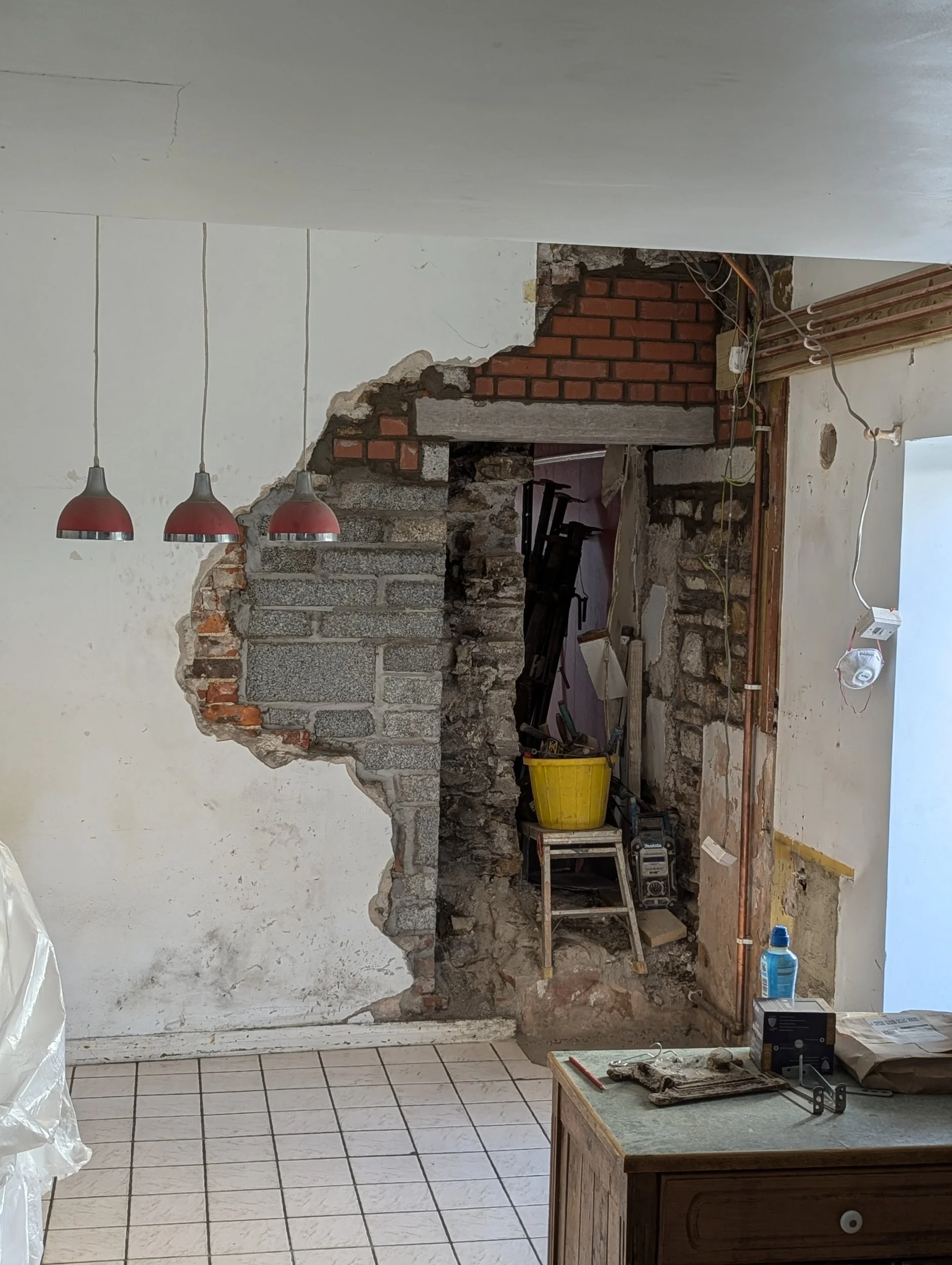Interior room under renovation with a partially demolished wall revealing brick and stone construction, construction tools, and supplies on a small table.