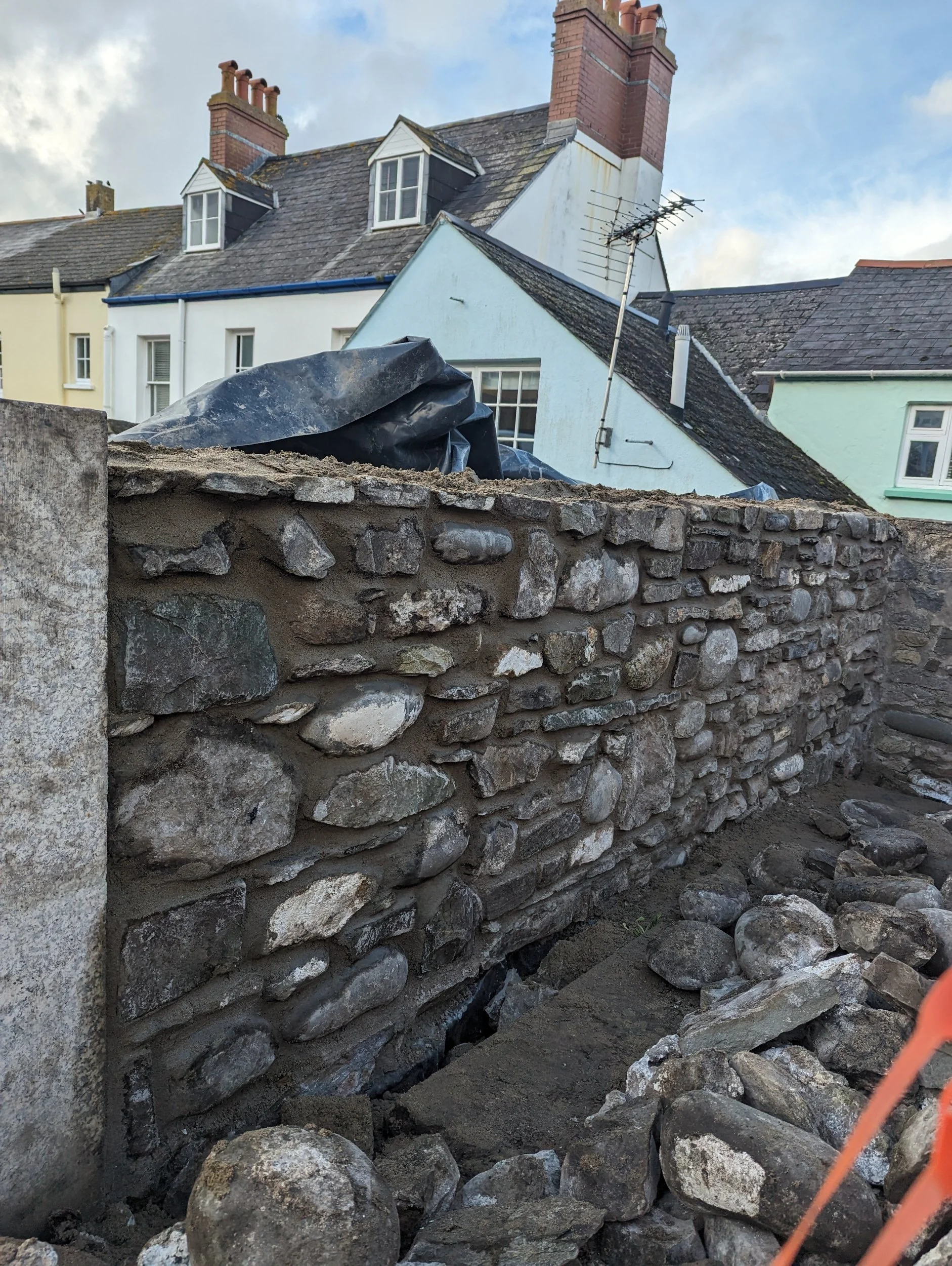 A stone wall under construction with various sizes of stones and mortar, set against a backdrop of residential houses with gabled roofs and chimneys.
