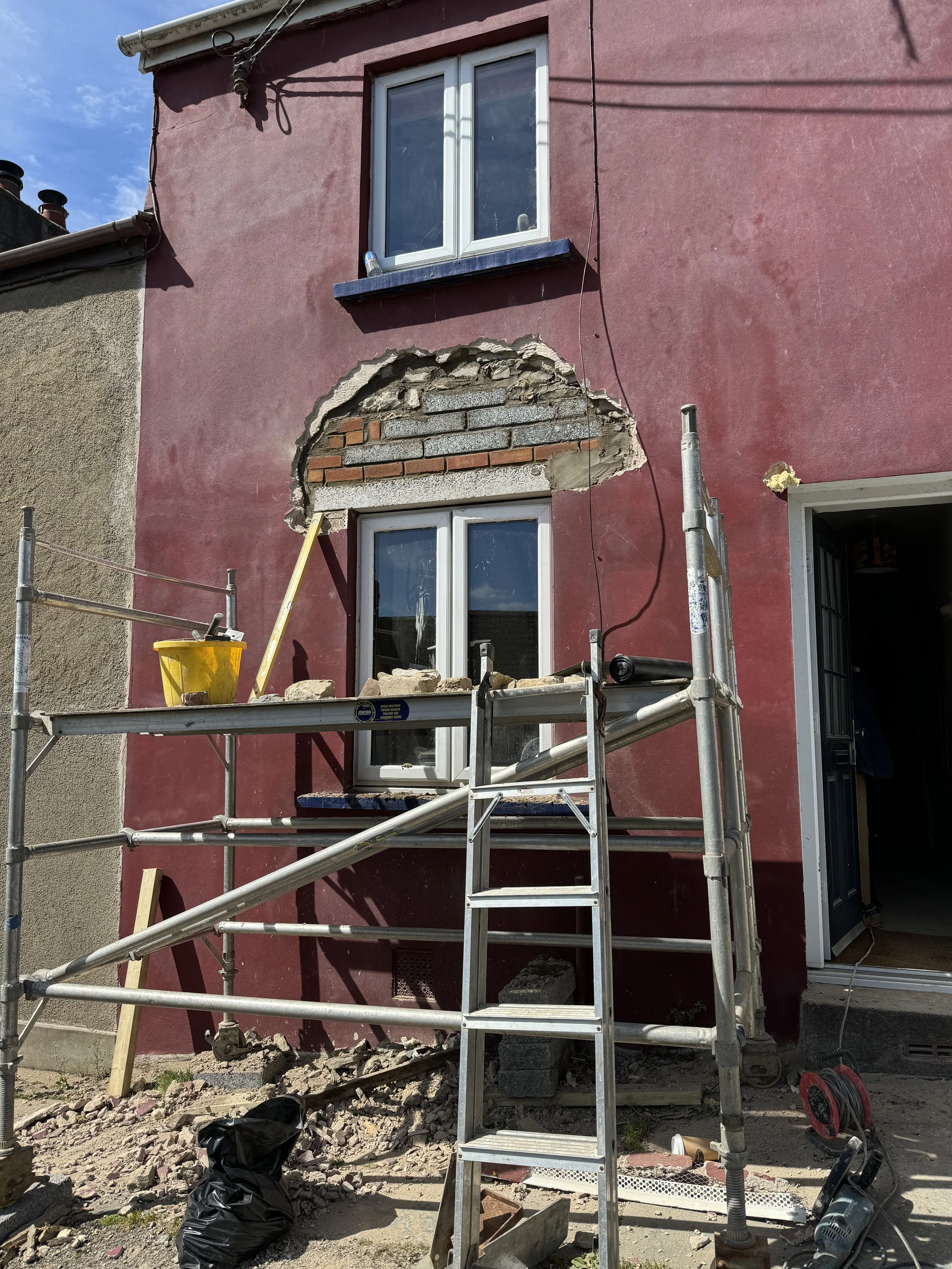 A building undergoing exterior renovation with scaffolding, exposing a section of brickwork and damaged wall beneath a window, with construction tools and materials nearby.