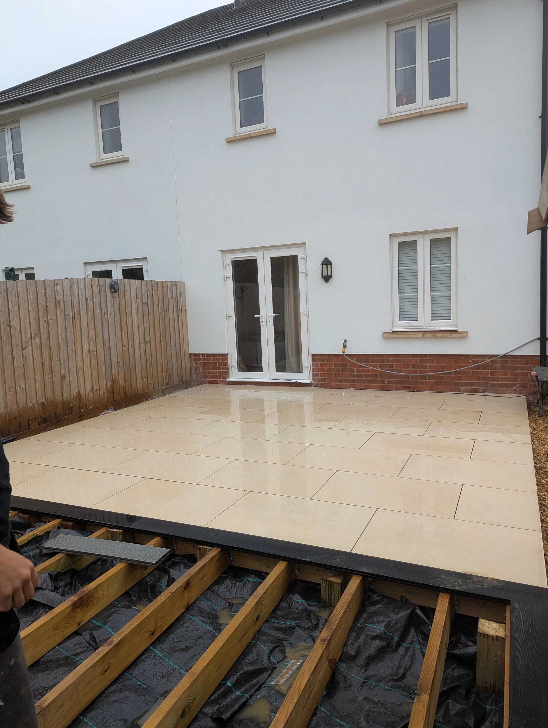 A newly installed stone patio in a backyard, with the remaining wooden frame of the deck visible in the foreground, rain droplets on the surface, a wooden fence on the left, and a white house with a sliding glass door and windows in the background.