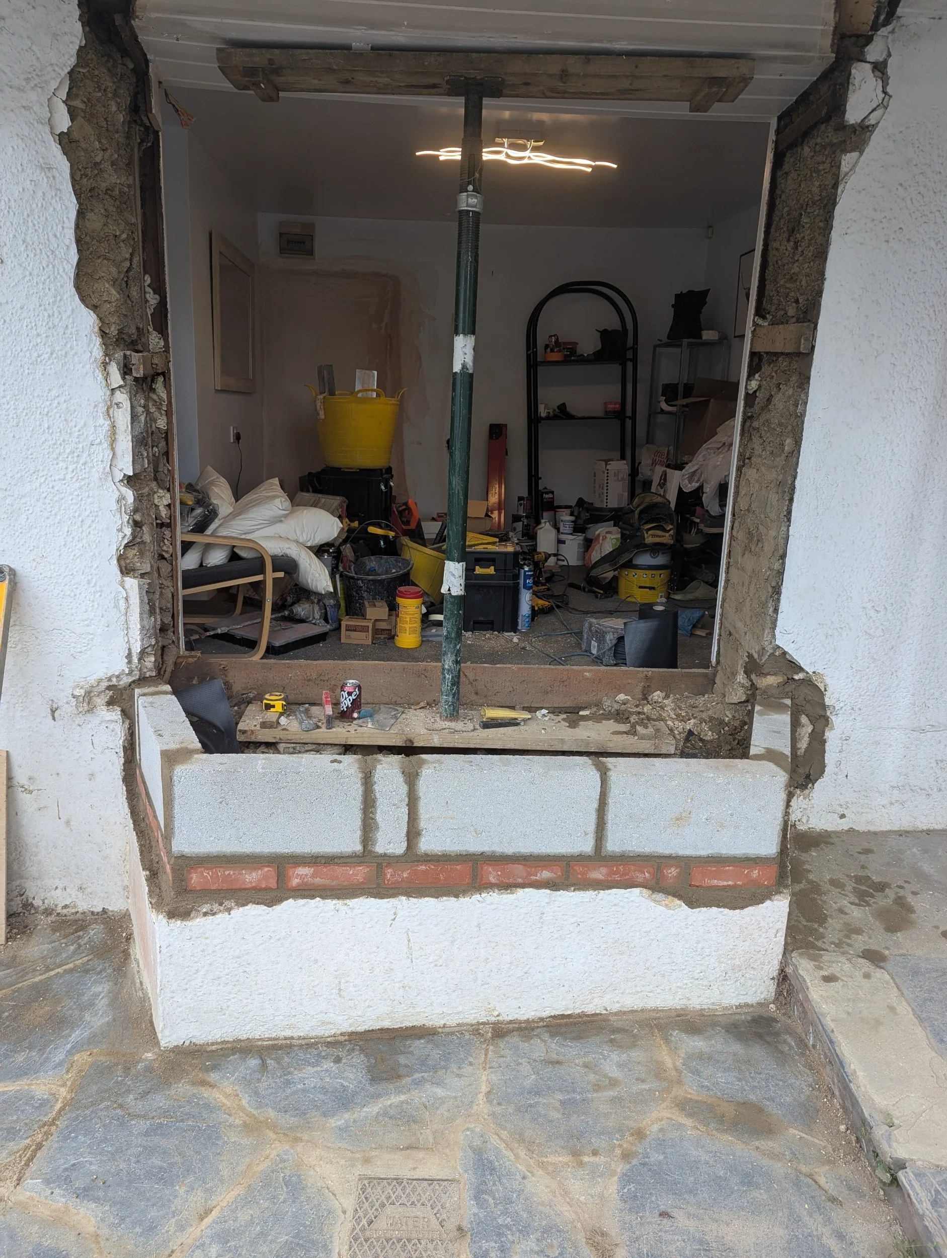 Photo shows a construction site with an opening in a wall, revealing a cluttered room filled with tools, boxes, buckets, and construction materials.