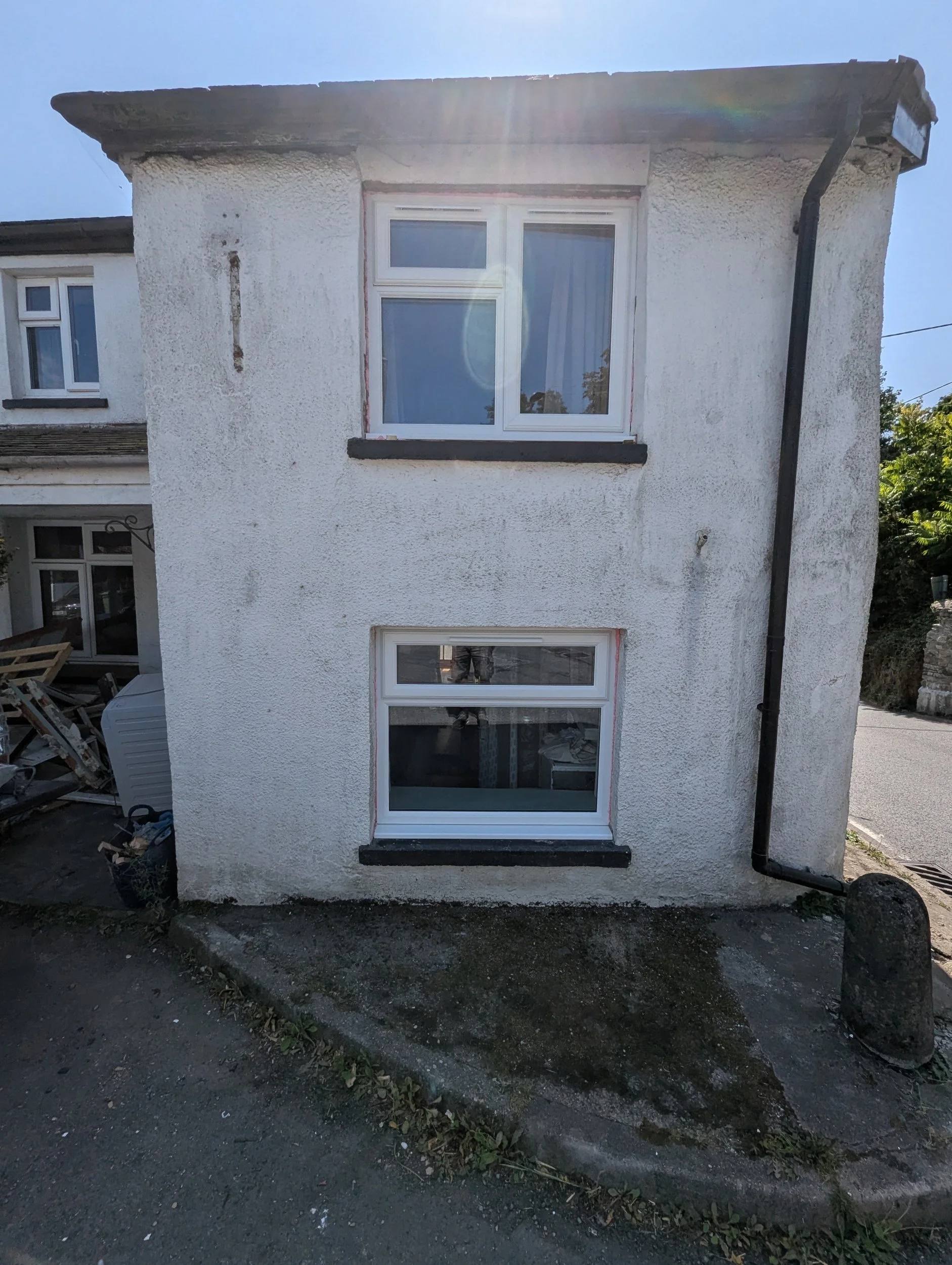 Two-story white house with textured exterior walls, two rectangular windows with white frames, black sills, and partially open blinds, on a sloped street with a sidewalk and drain, and trees in the background.