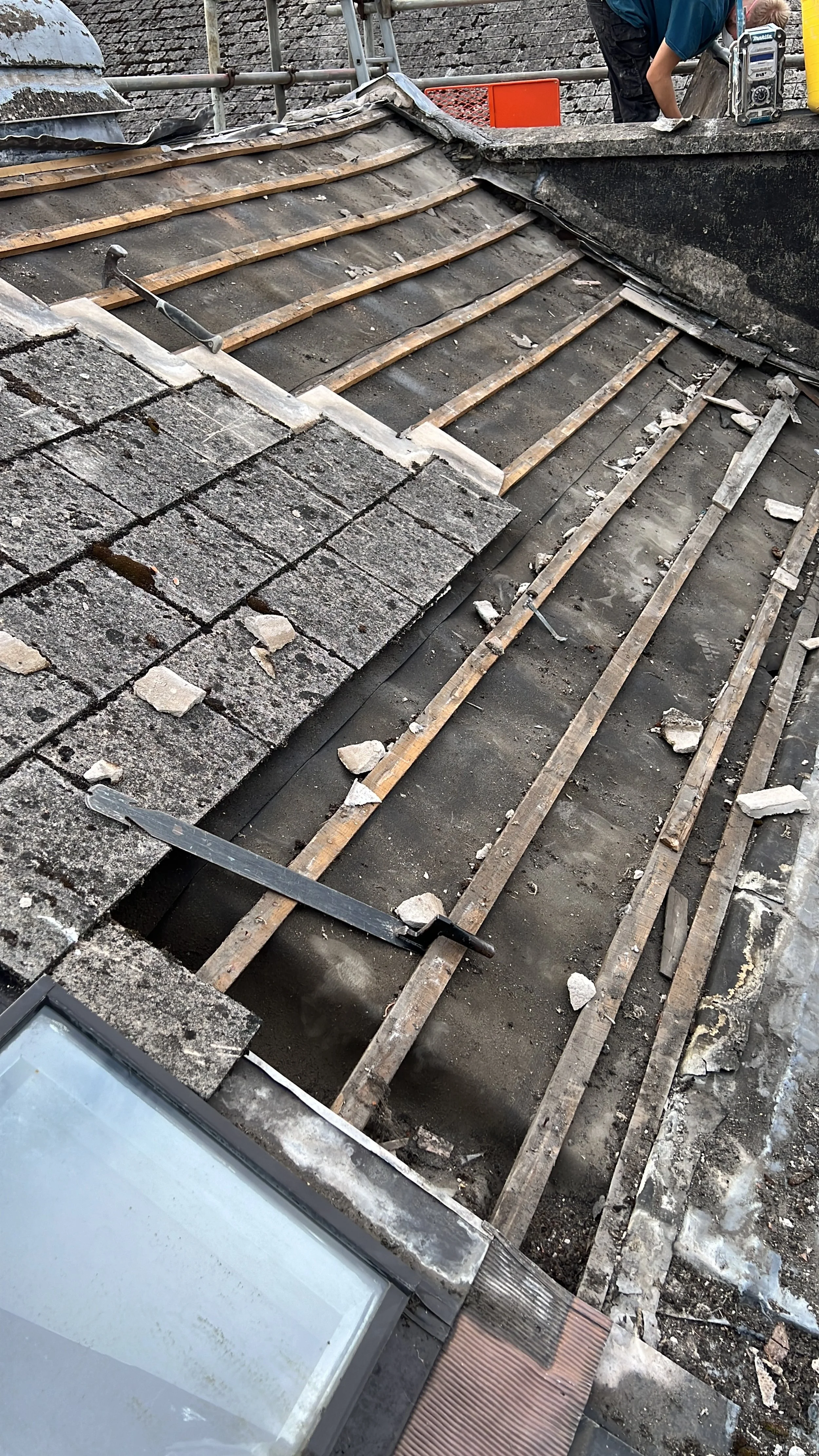 Roof undergoing repair with removed shingles, exposing wooden battens and underlayment, with a worker in the background and tools visible.