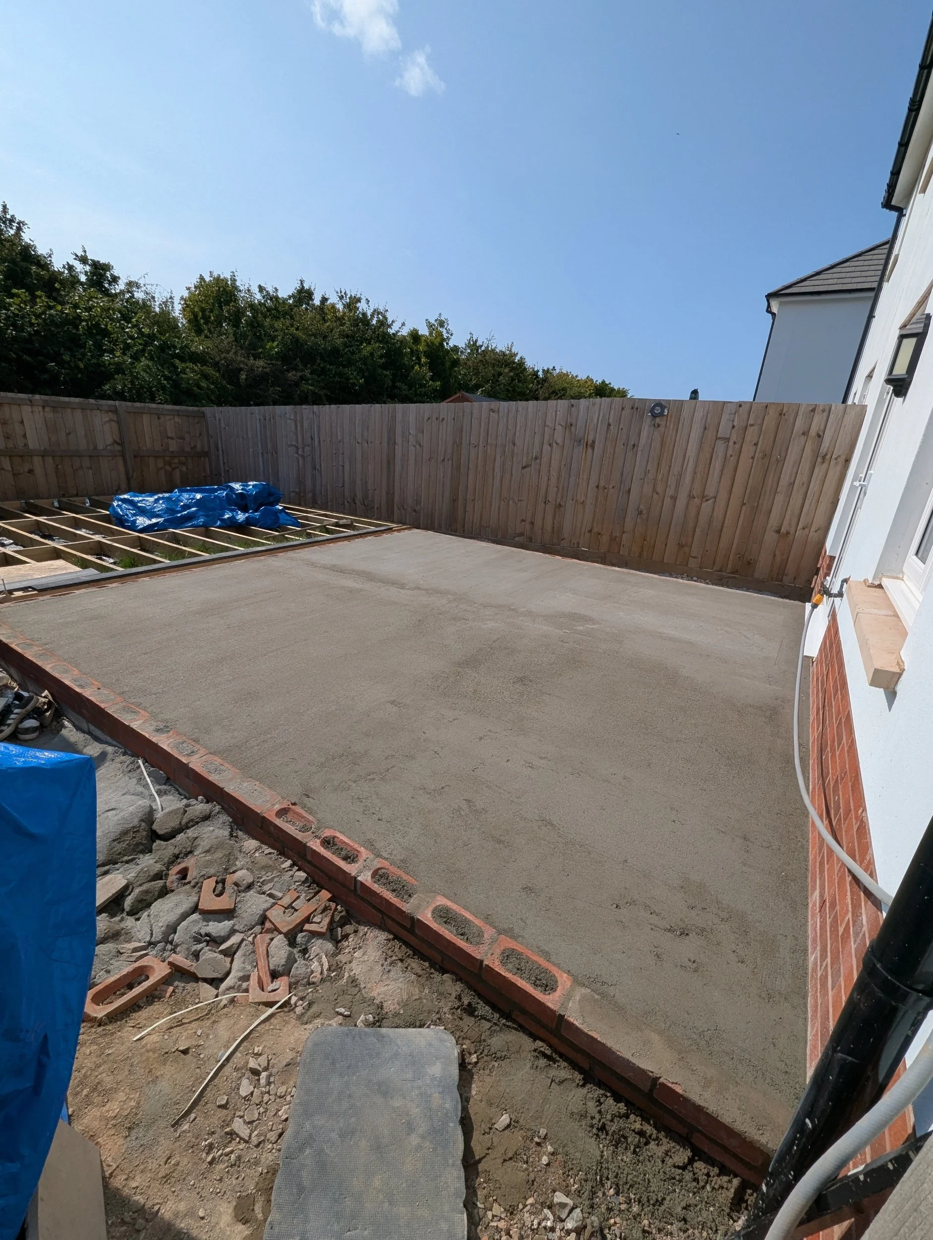 Newly poured concrete patio in a backyard with a wooden fence and a white house, under a blue sky.