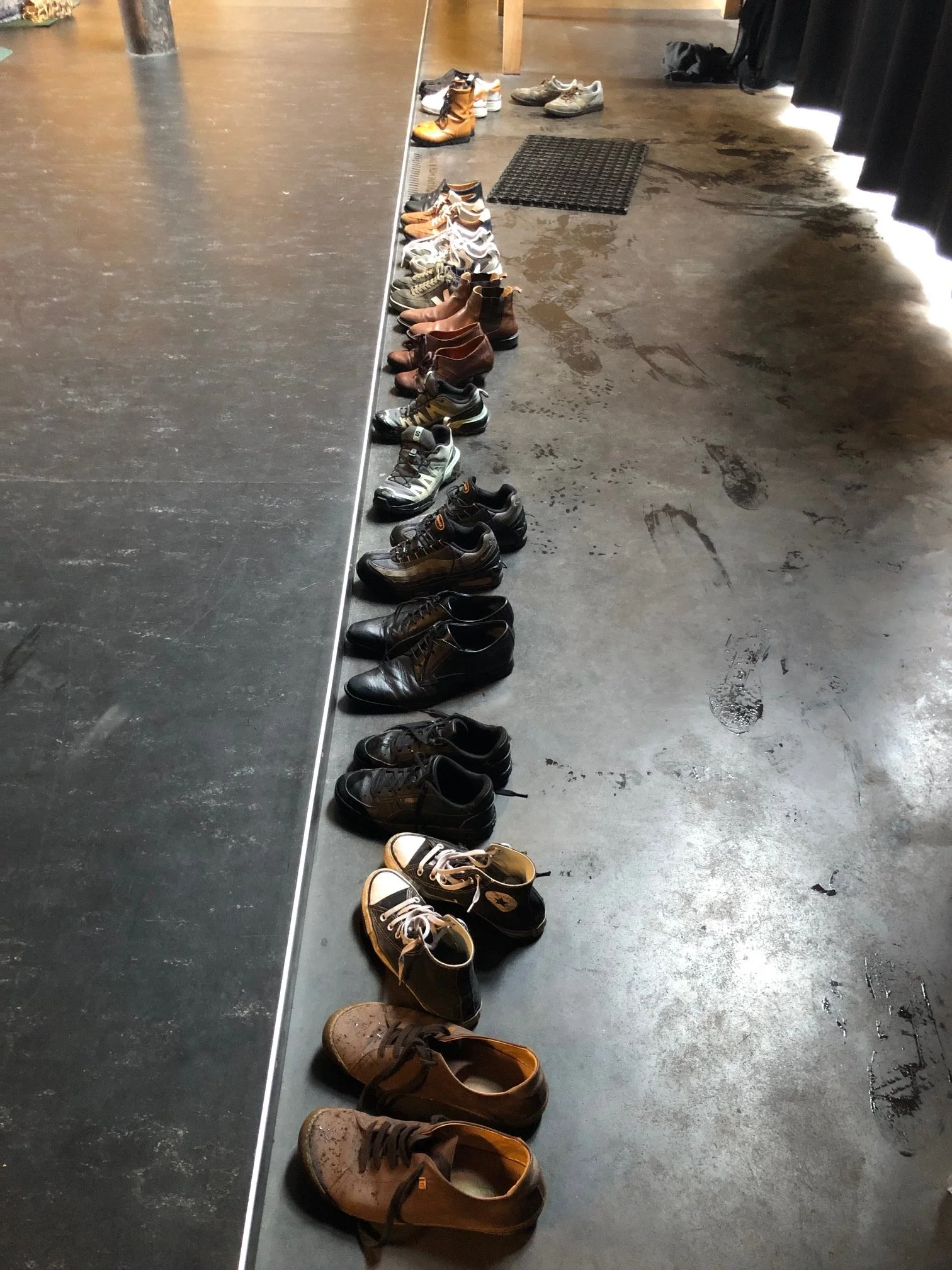 Rijen schoenen en laarzen op de vloer naast een muur, mogelijk in een hal of kleedruimte.